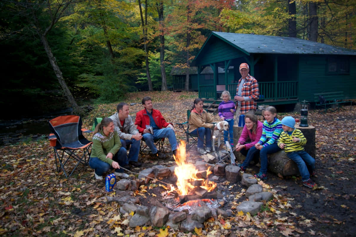 Fall Camping 桌布 壁紙 — 一家人圍著噼啪作響的營火，背景是秋葉森林和綠色小屋。