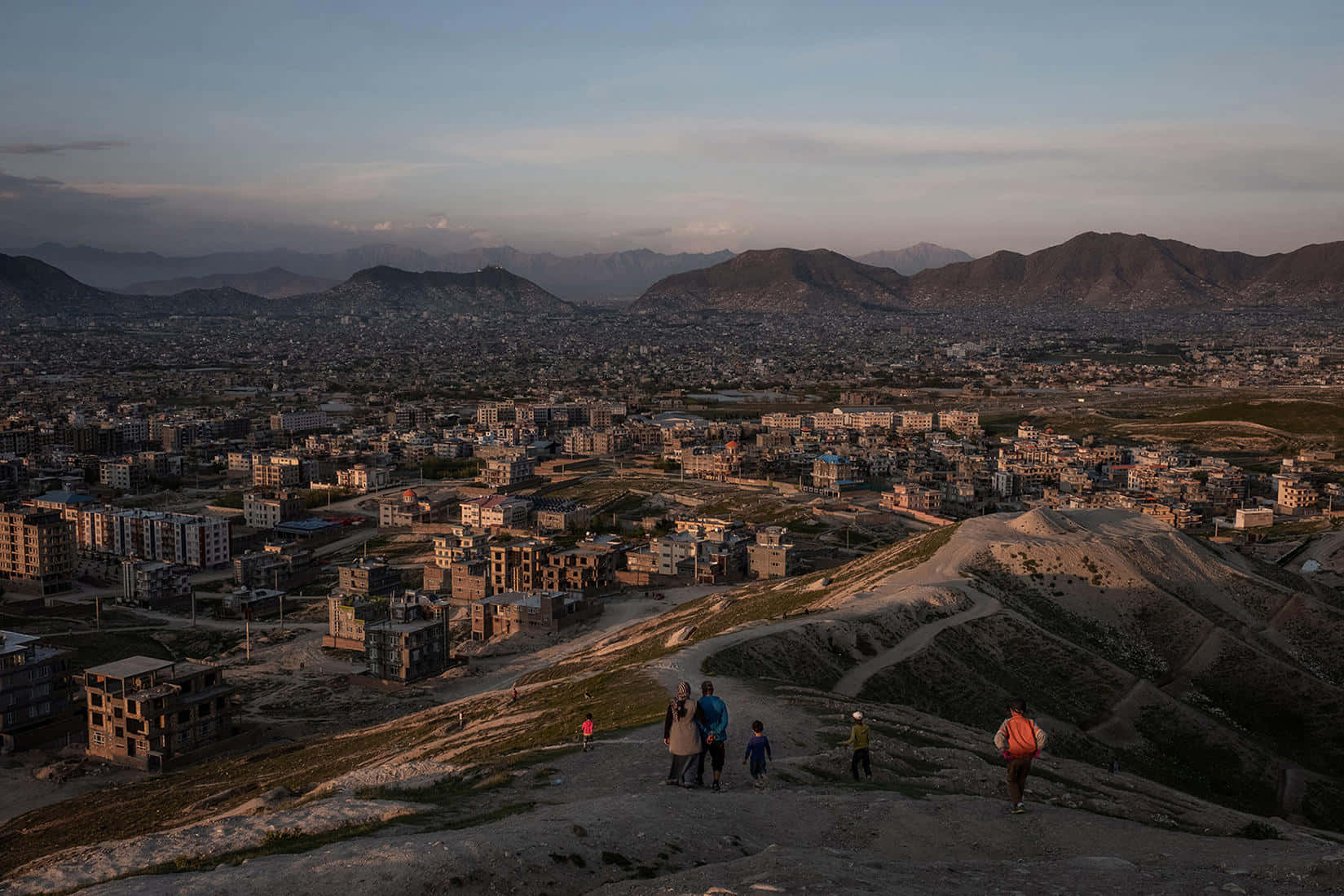 Afghanistan 桌布 壁紙 — 一家人在日落時分沿著塵土飛揚的山坡行走，俯瞰著坐落在遠山之間的廣闊城市。