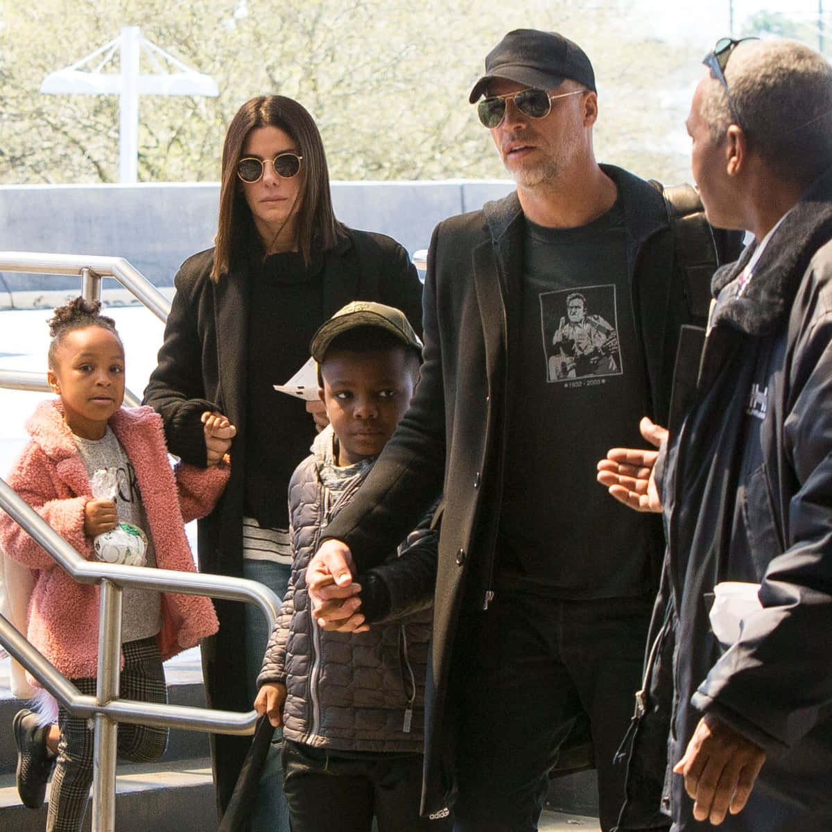 Sandra Bullock 2021 桌布 壁紙 — 一家人在明亮的日光下手牽手走下戶外樓梯，背景有樹木和白色建築物。