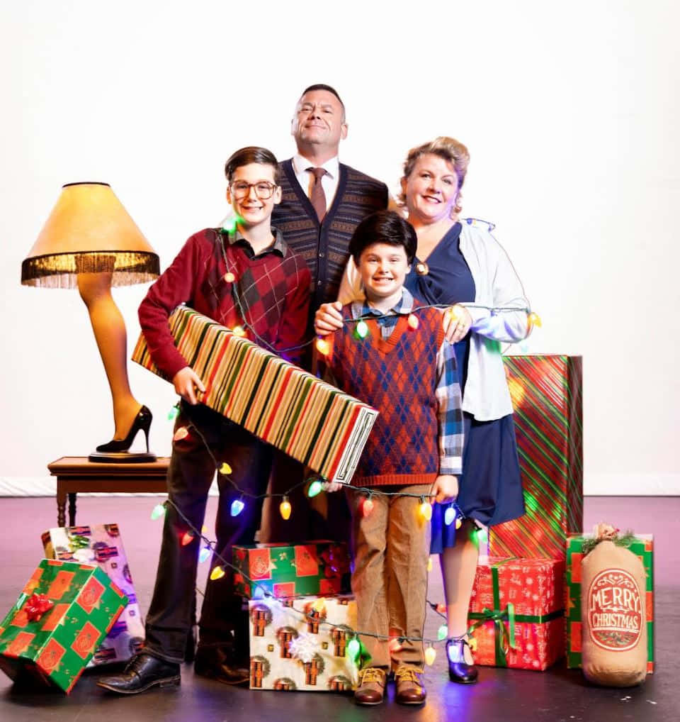 a christmas story 桌布 壁紙 — 一家人愉快地擺姿勢，拿著超大的聖誕禮物和節日燈飾，圍繞著桌上的燈。
