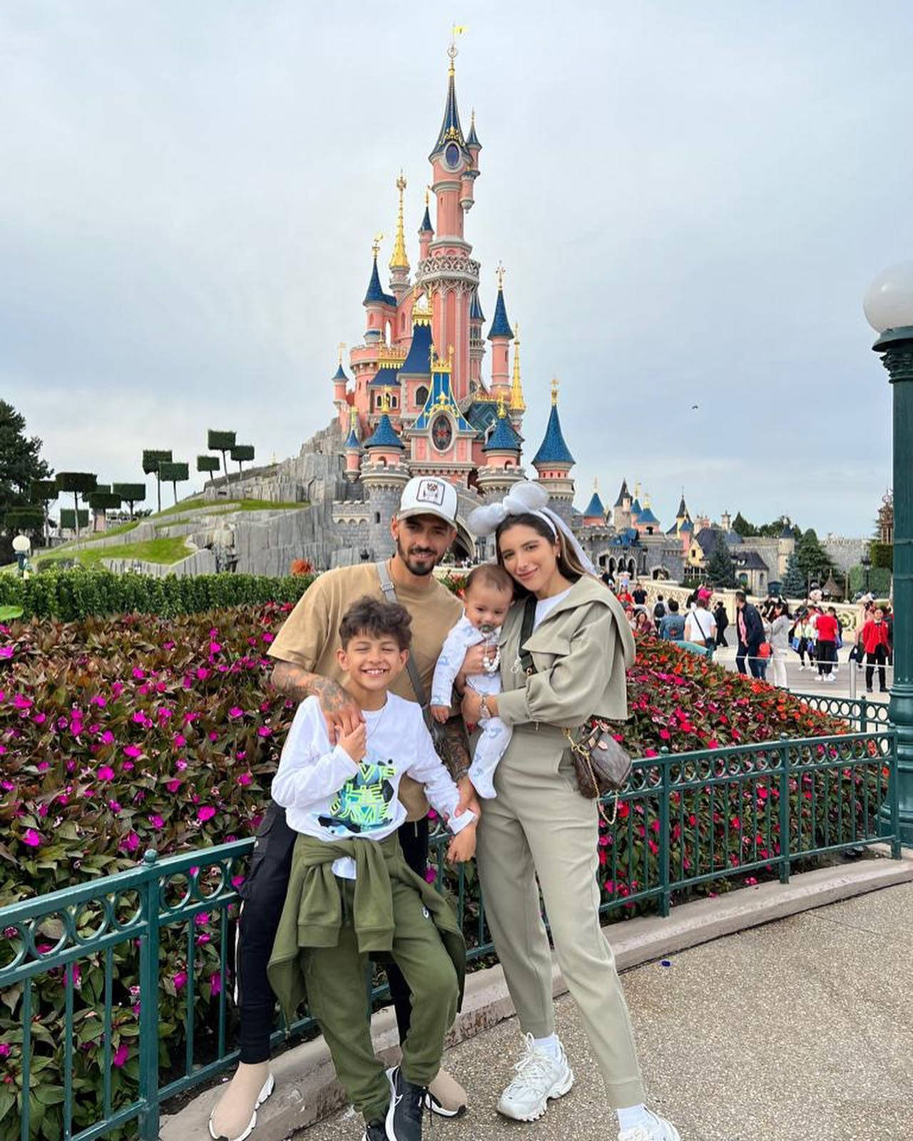 Disneyland Castle 桌布 壁紙 — 一家四口在標誌性的粉藍色迪士尼城堡前，在鮮豔的粉紅色花朵襯托下微笑拍照。