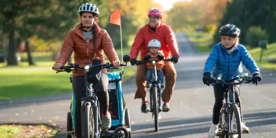 bike riding 桌布 壁紙 — 一家四口在溫暖的秋日陽光下，沿著綠樹成蔭的小徑騎腳踏車。