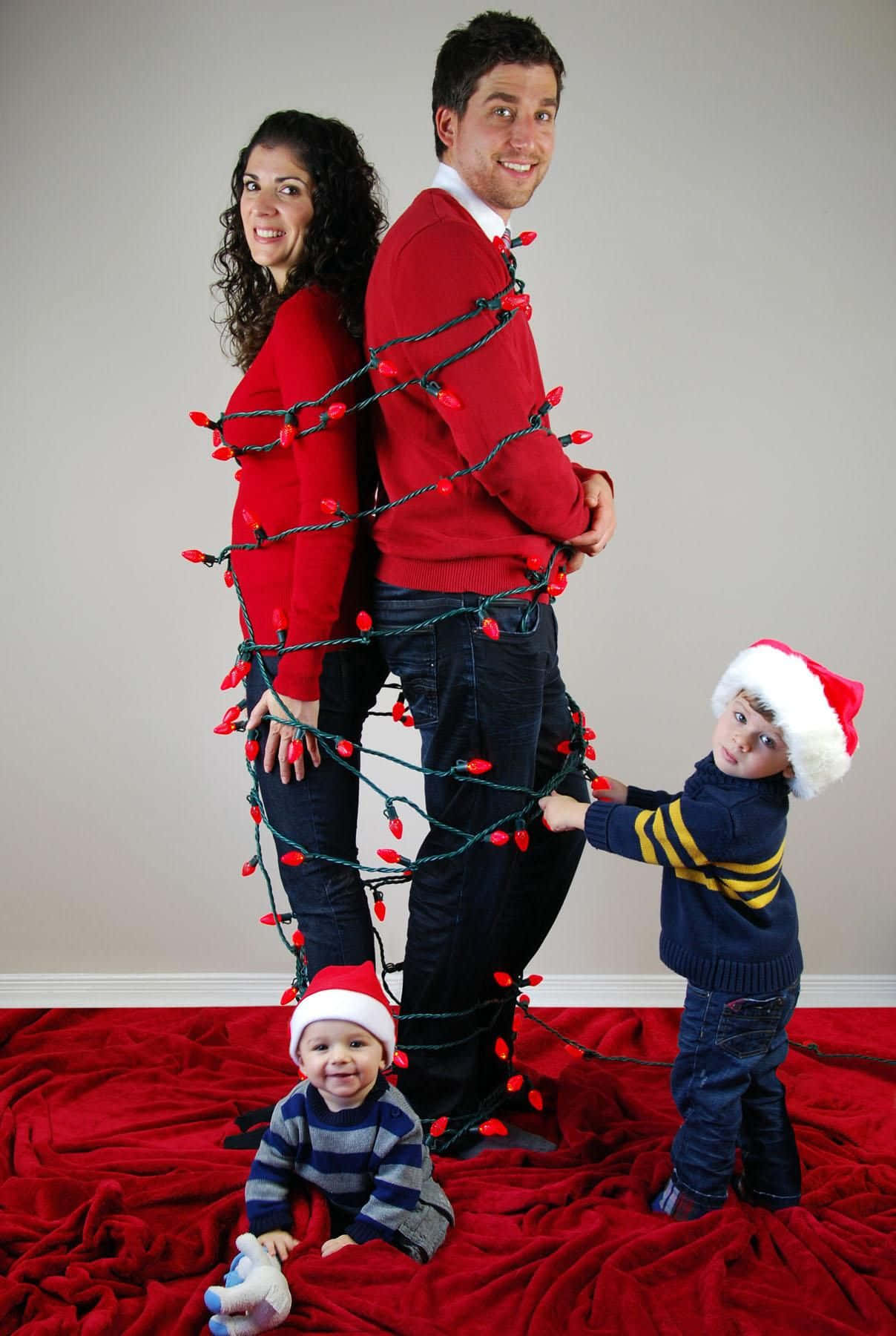 Family Christmas 桌布 壁紙 — 一家四口在紅色地毯上，對著素色牆壁，玩味十足地用紅色聖誕燈飾纏繞身體擺姿勢。
