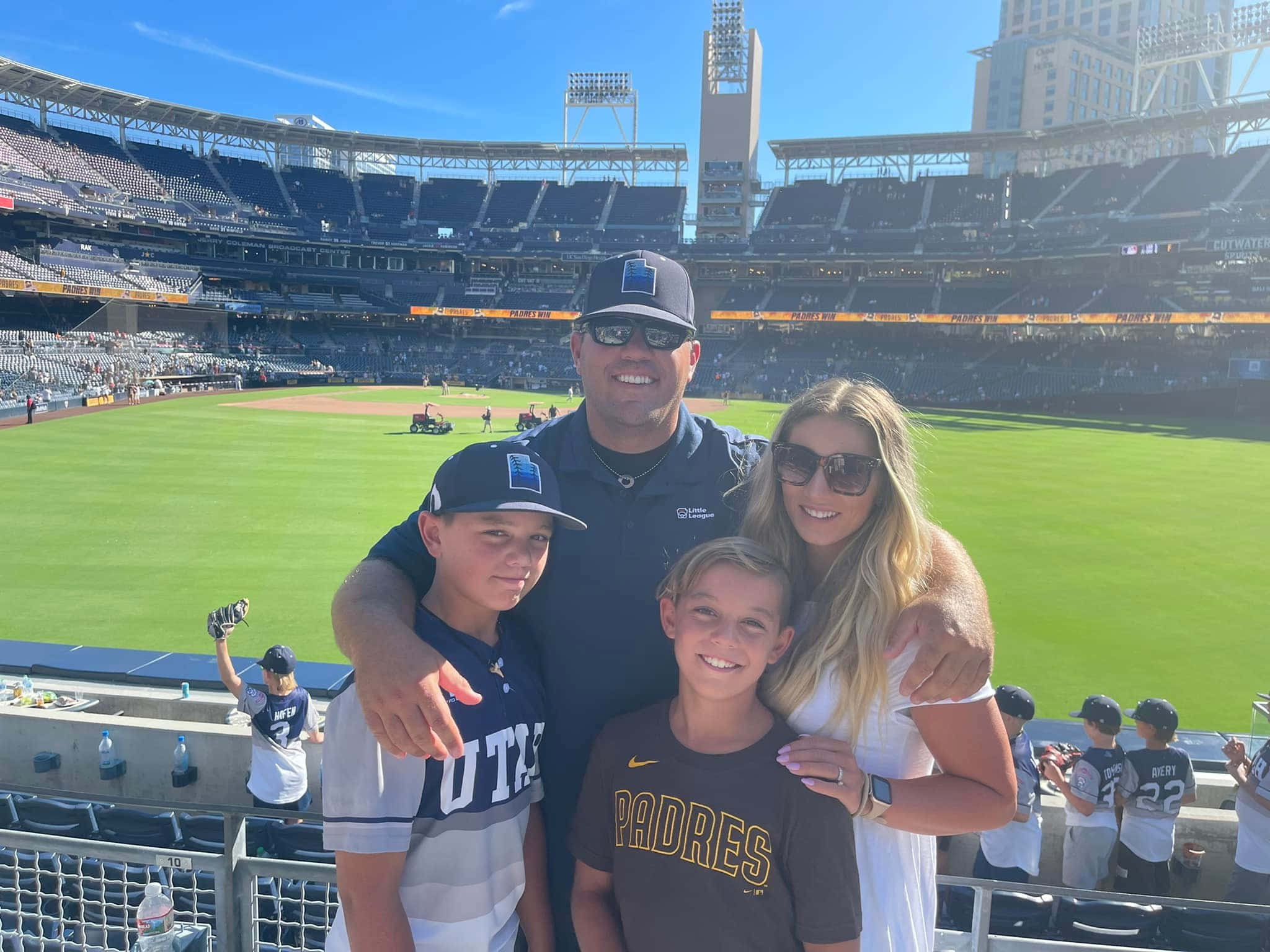 Little League Baseball 桌布 壁紙 — 一家四口在陽光明媚的棒球場上微笑拍照，身後是綠色球場和空無一人的看台。
