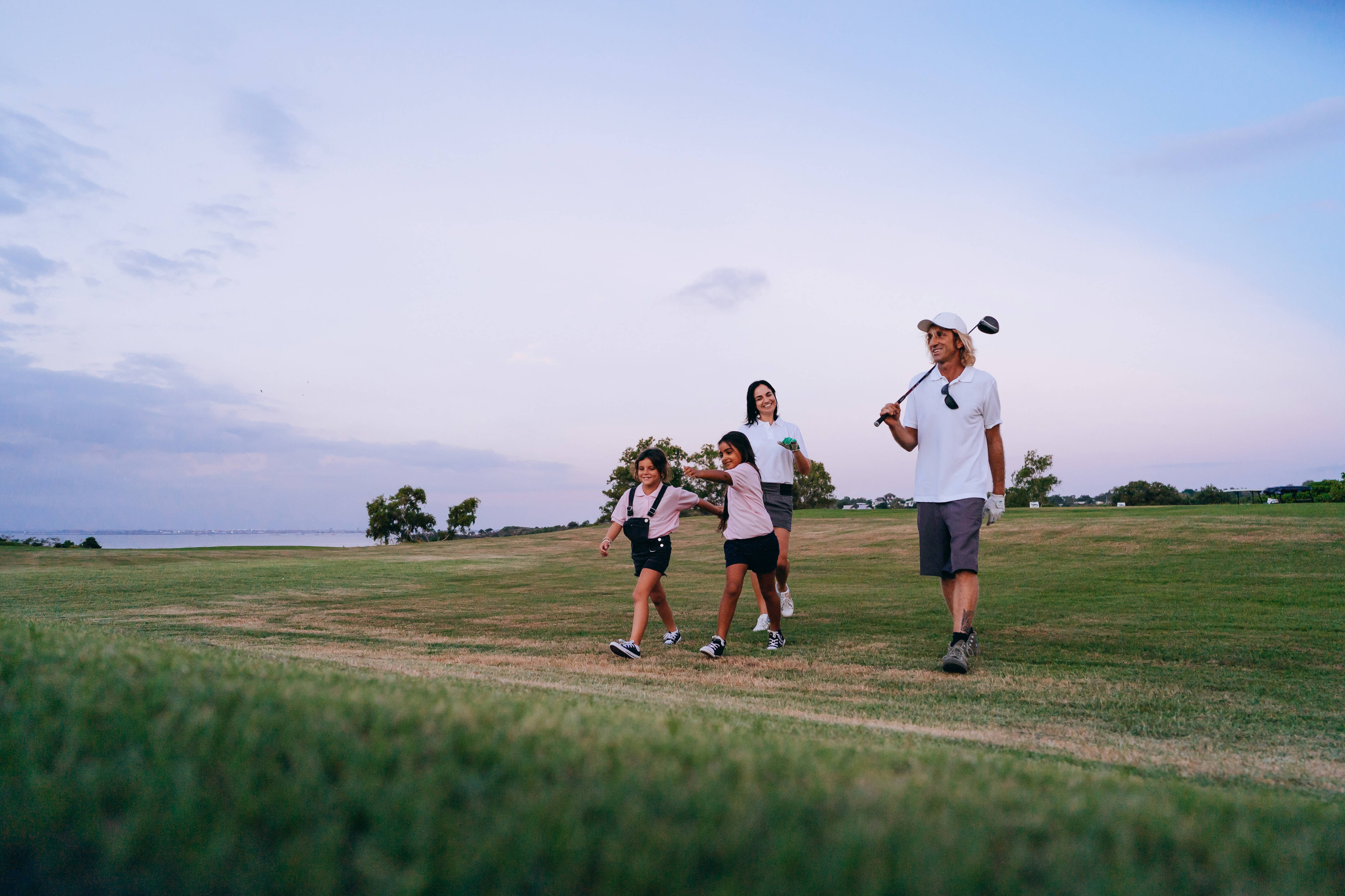 4K Golf 桌布 壁紙 — 一家四口在黃昏時分走過草地高爾夫球場，父親拿著高爾夫球桿，孩子們在他身邊大笑。
