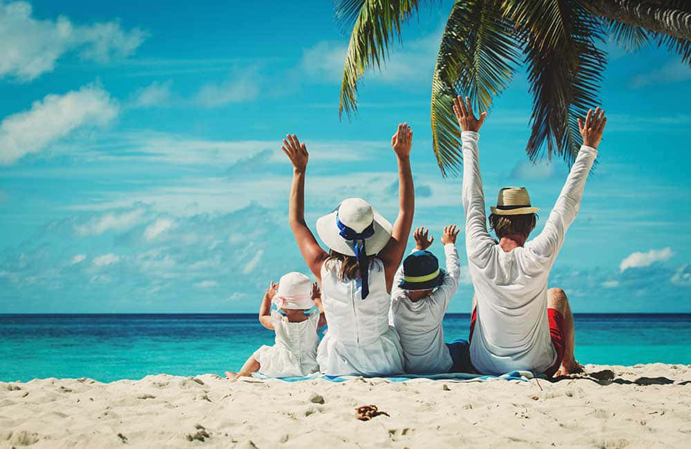 Beach Family 桌布 壁紙 — 一家四口坐在熱帶海灘上，舉起雙臂朝向頭頂棕櫚葉映襯的明亮藍天。