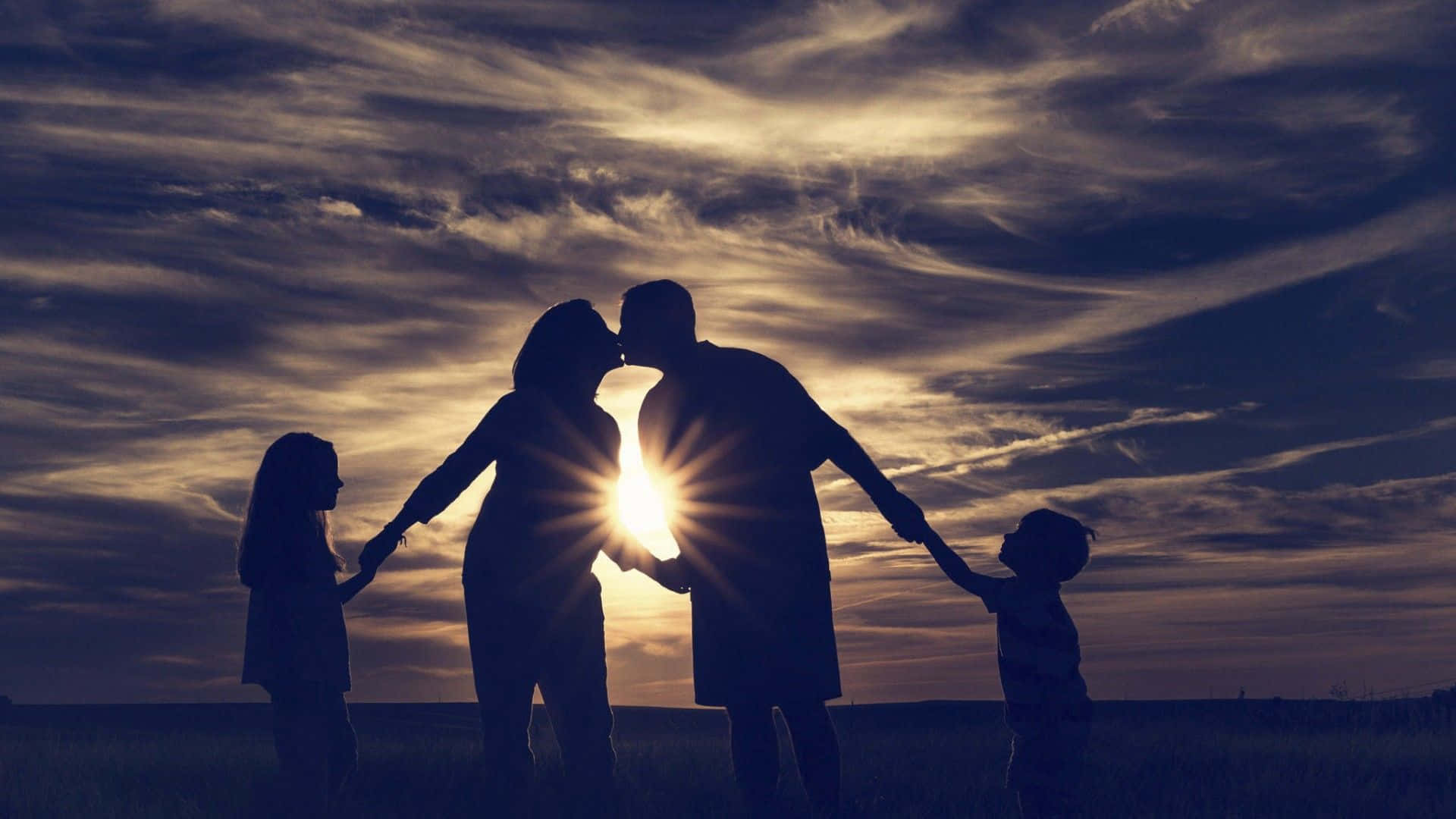Family 桌布 壁紙 — 一家四口手牽手，以戲劇性的日落天空為背景形成剪影，背後有燦爛的太陽光芒。