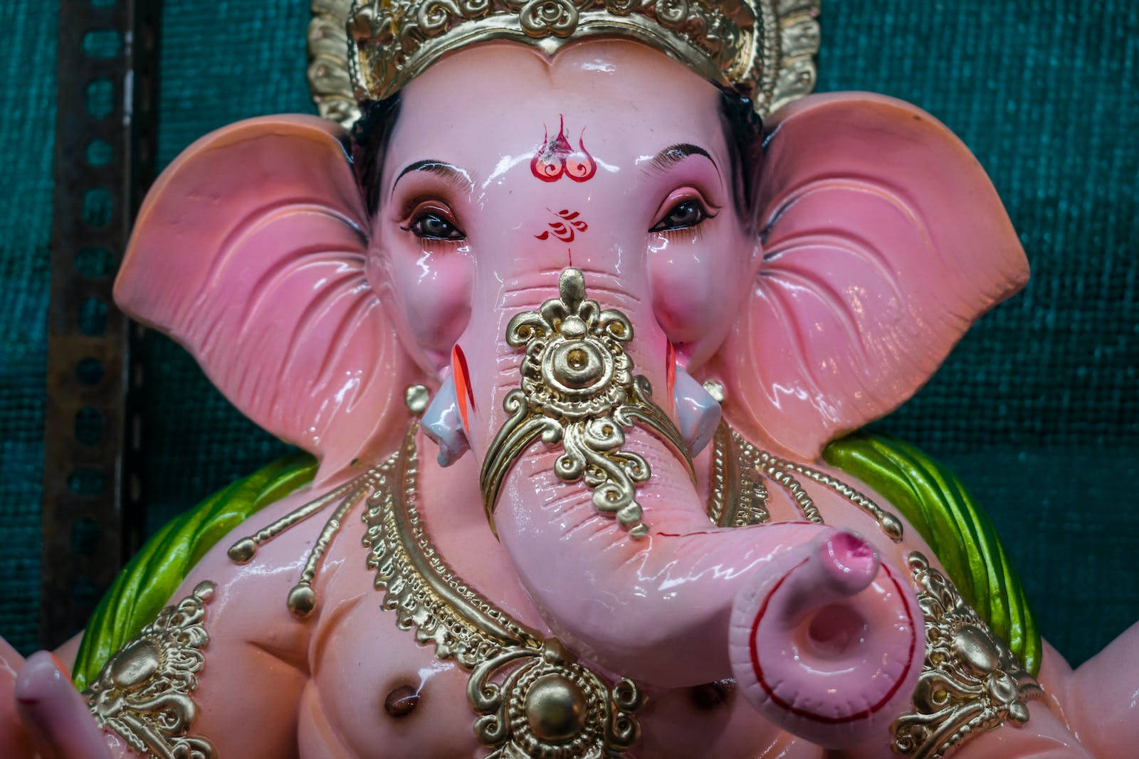Vinayaka 桌布 壁紙 — 一尊亮粉色的象神雕像，飾有金色飾物和紅色標記，背景為青綠色布料。
