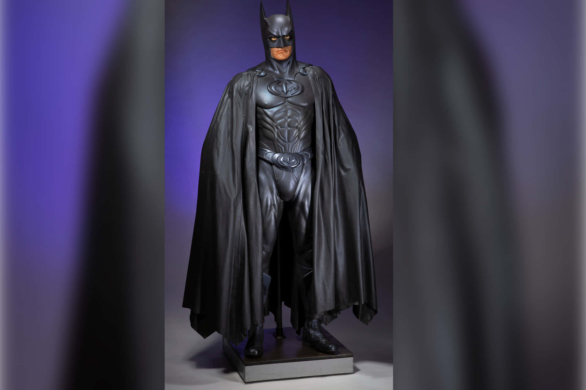 Bat-suit 桌布 壁紙 — 一尊亮黑色蝙蝠俠雕像矗立在基座上，背景為紫色漸層，帶有戲劇性的燈光效果。