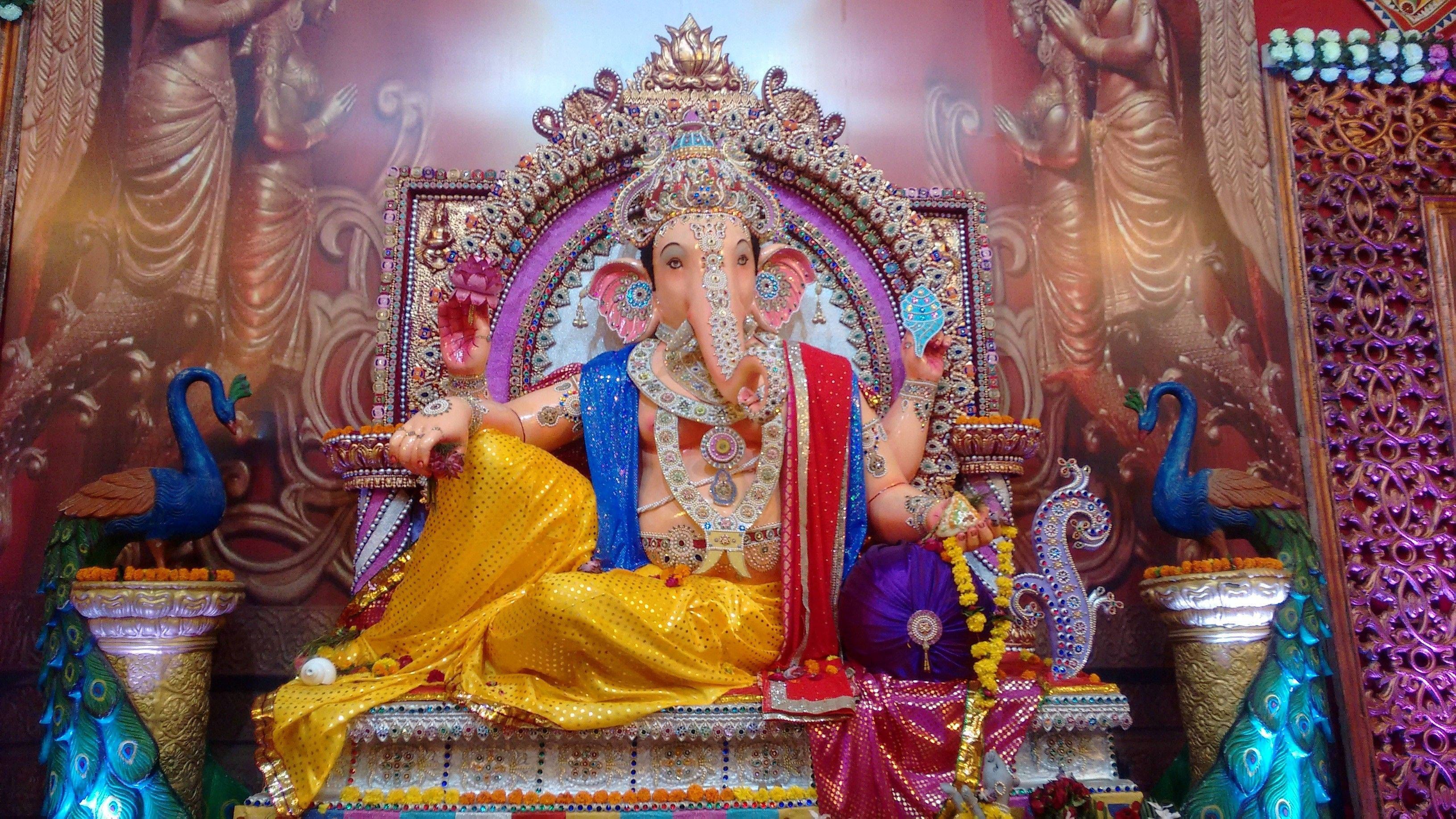 Hindu God 桌布 壁紙 — 一尊以金色、藍色和紅色裝飾的象神雕像，威嚴地坐在華麗的寶座上，兩側有孔雀相伴。