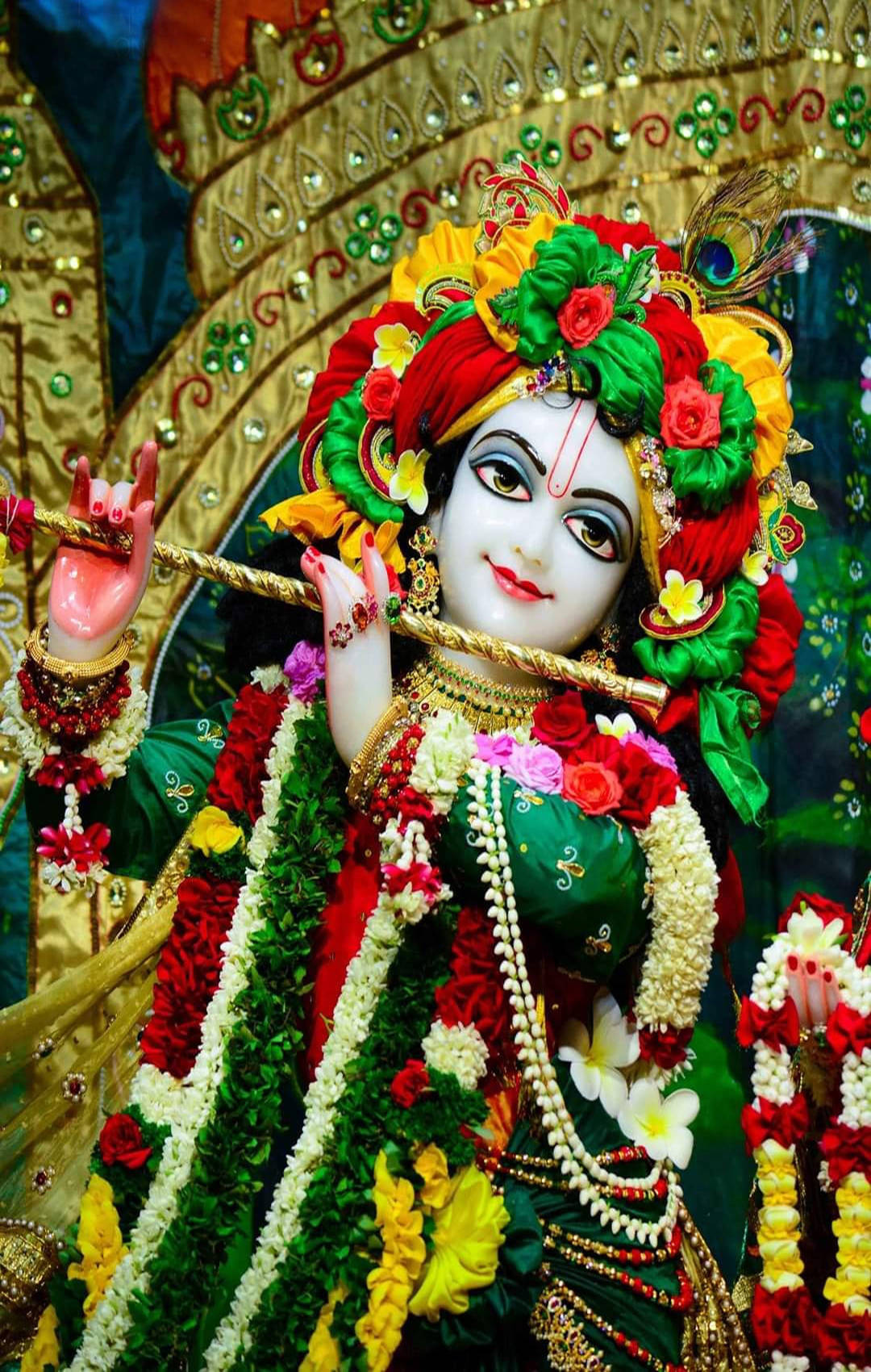 krishna iPhone 桌布 壁紙 — 一尊充滿活力的克里希納神雕像，飾有色彩繽紛的花朵、珠寶和孔雀羽毛，在金色華麗的背景前手持長笛。