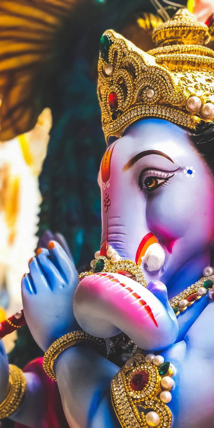 cute ganesha 桌布 壁紙 — 一尊充滿活力的藍色象神雕像，飾有金色珠寶和彩色寶石，象鼻中拿著粉紅色的糖果。