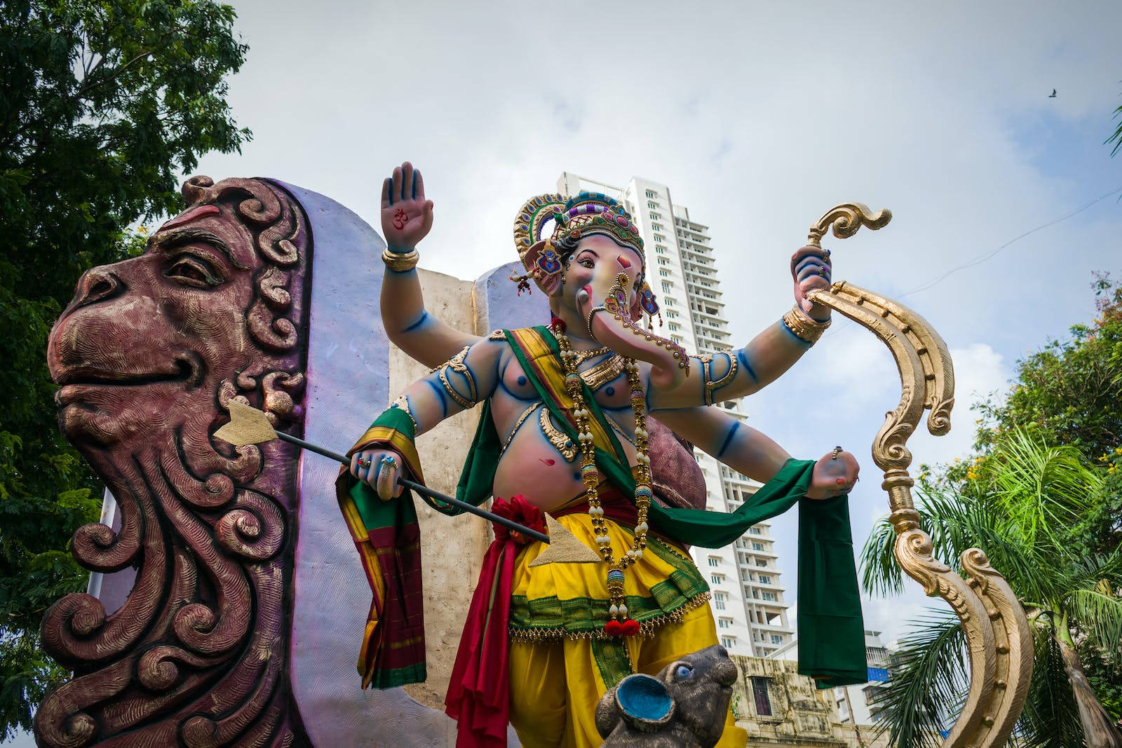 Ganesh 桌布 壁紙 — 一尊充滿活力的象頭神像，在現代摩天大樓和茂密綠植的背景下，勝利地斬下一條巨蛇的頭。
