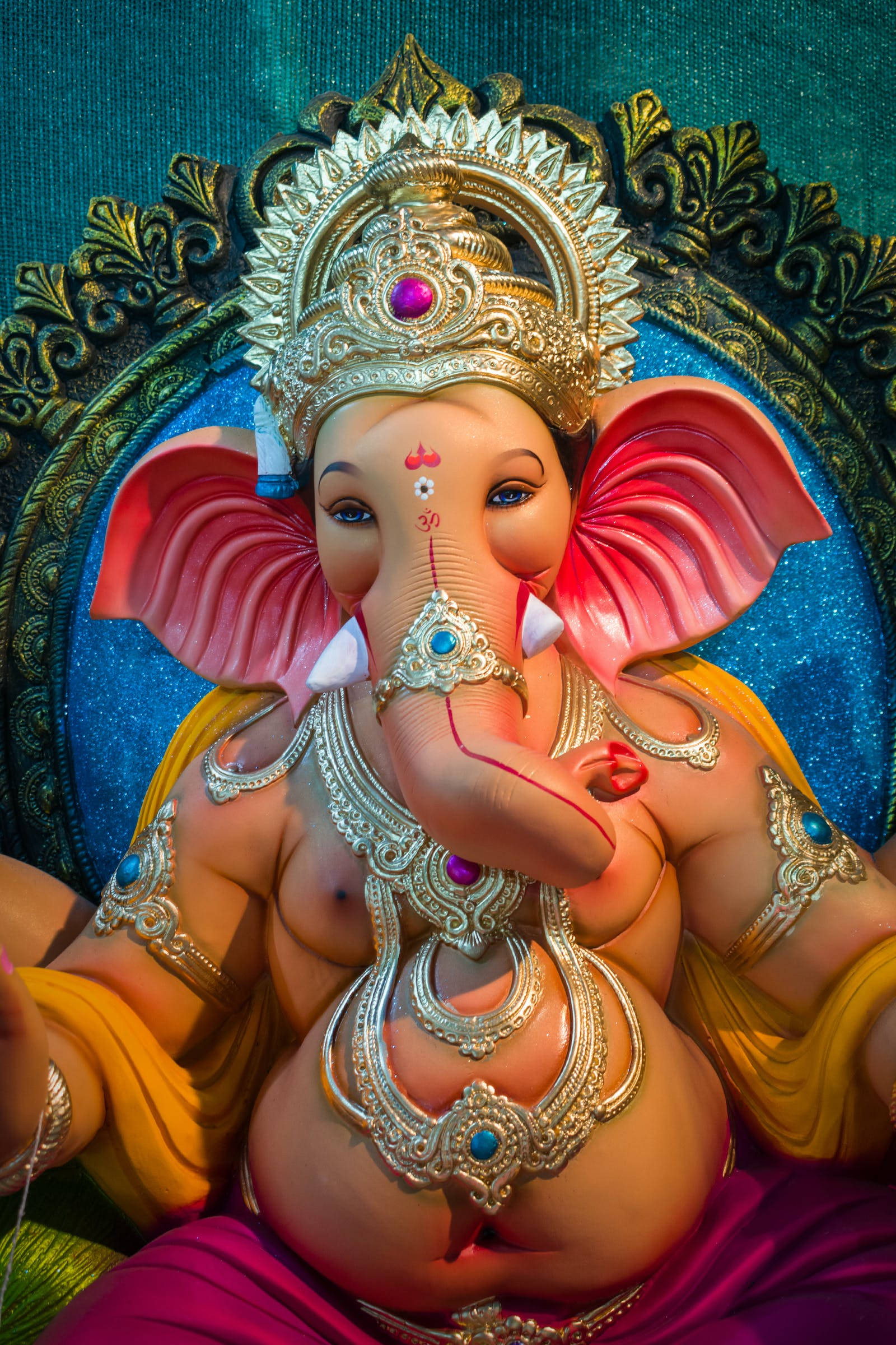 Vinayaka 桌布 壁紙 — 一尊充滿活力的象頭神像，飾有金色珠寶和粉紅色象頭，坐在藍綠色和金色的華麗背景前。