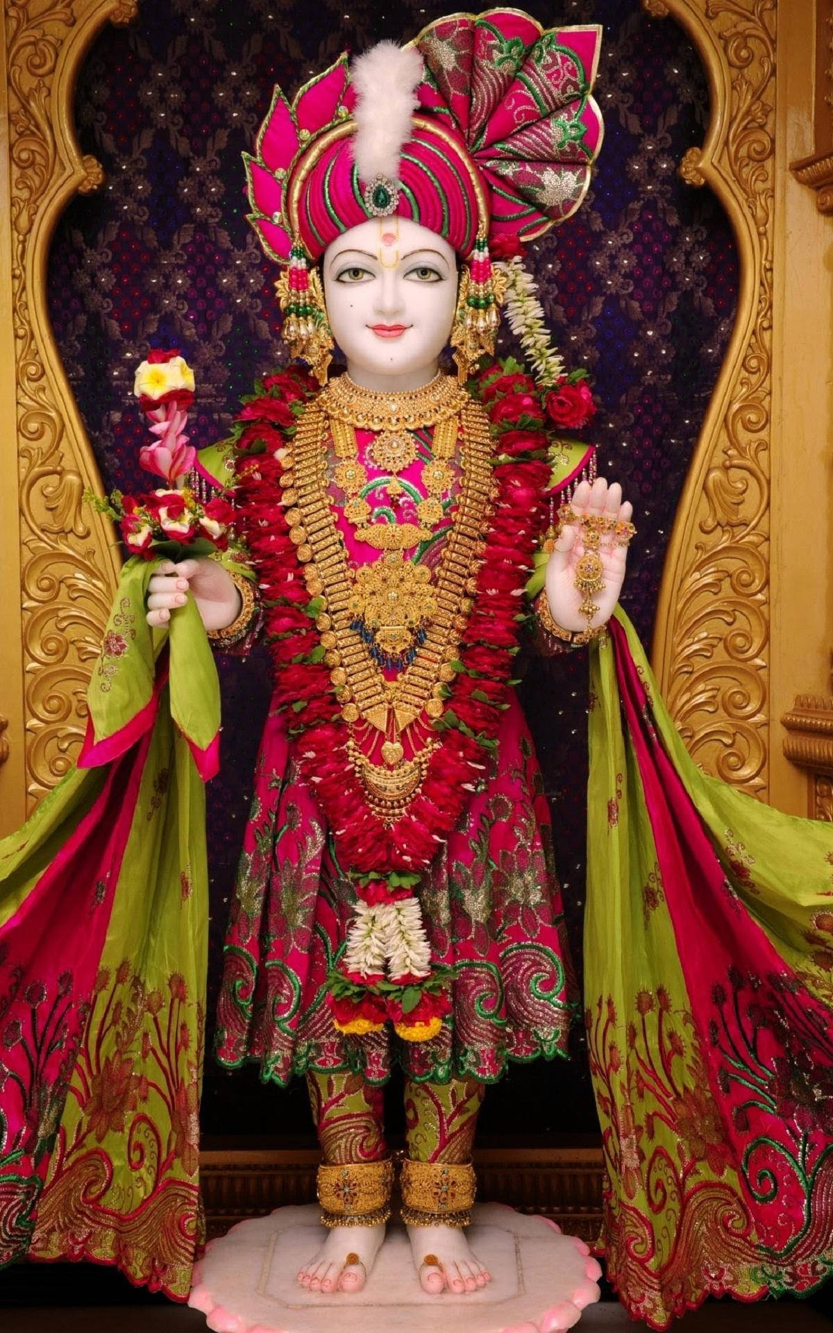 Swaminarayan 桌布 壁紙 — 一尊光彩奪目的神像，身著鮮豔的粉紅色和綠色服飾，佩戴金色珠寶，優雅地站在華麗的金色拱門前。