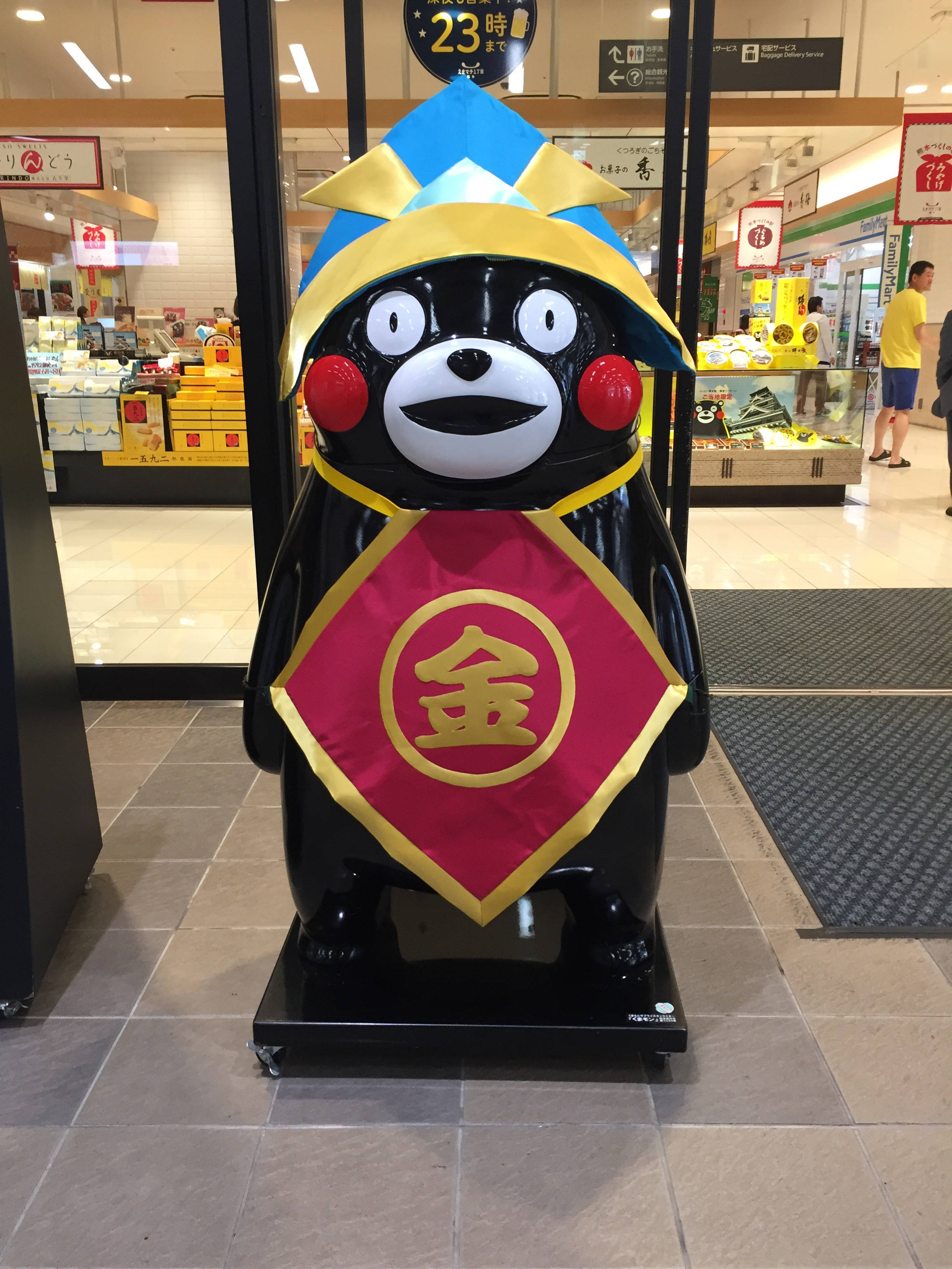 熊本熊 桌布 壁紙 — 一尊光滑的黑色熊本熊雕像，戴著藍黃色帽子和帶有漢字的紅金色腰帶，站在光線明亮的商店通道中。