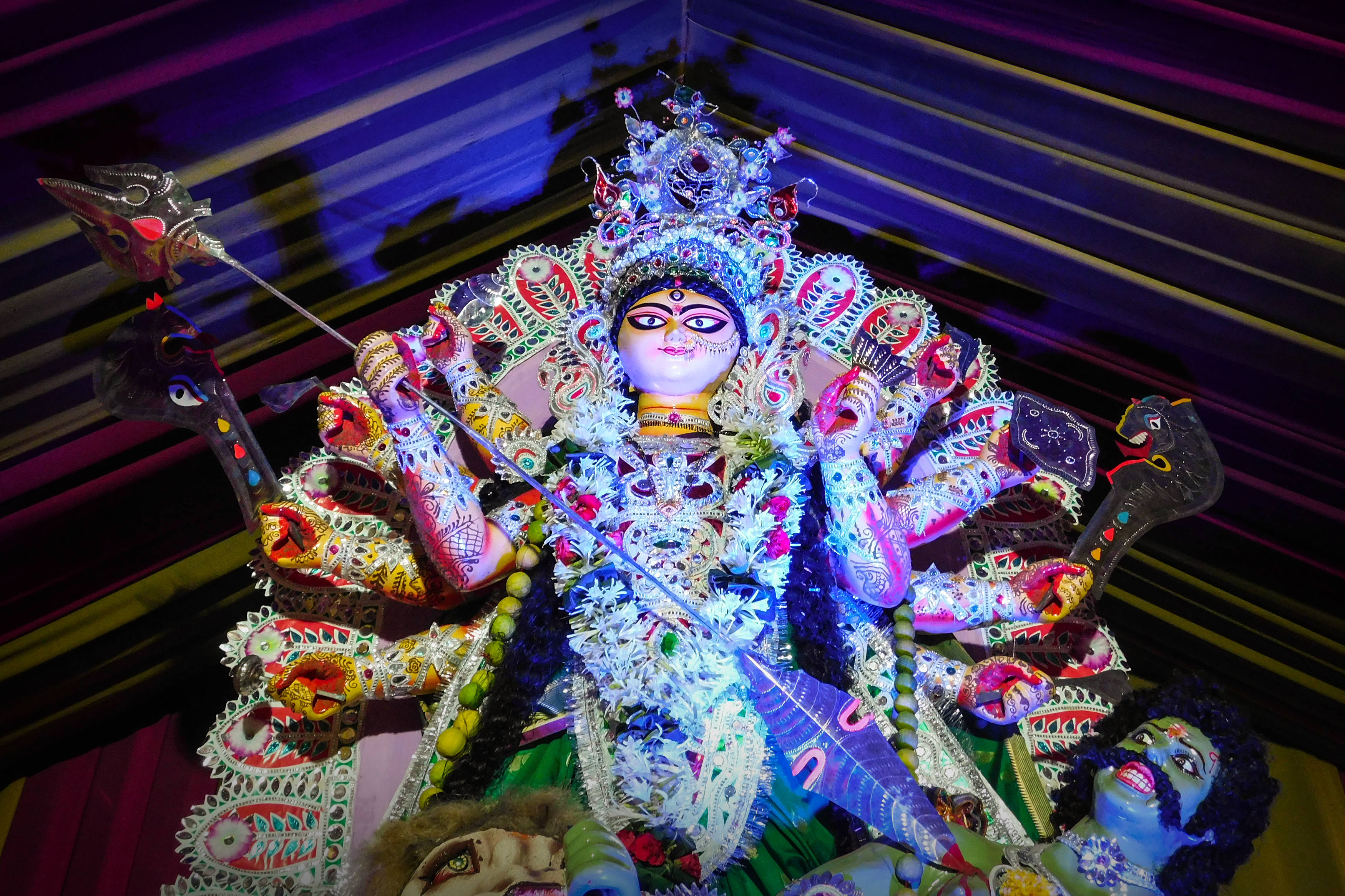 Navratri 桌布 壁紙 — 一尊光芒四射、多臂的杜爾迦神像，飾有精緻的珠寶和武器，在條紋背景襯托下，被鮮豔的紫色和藍色燈光照亮。