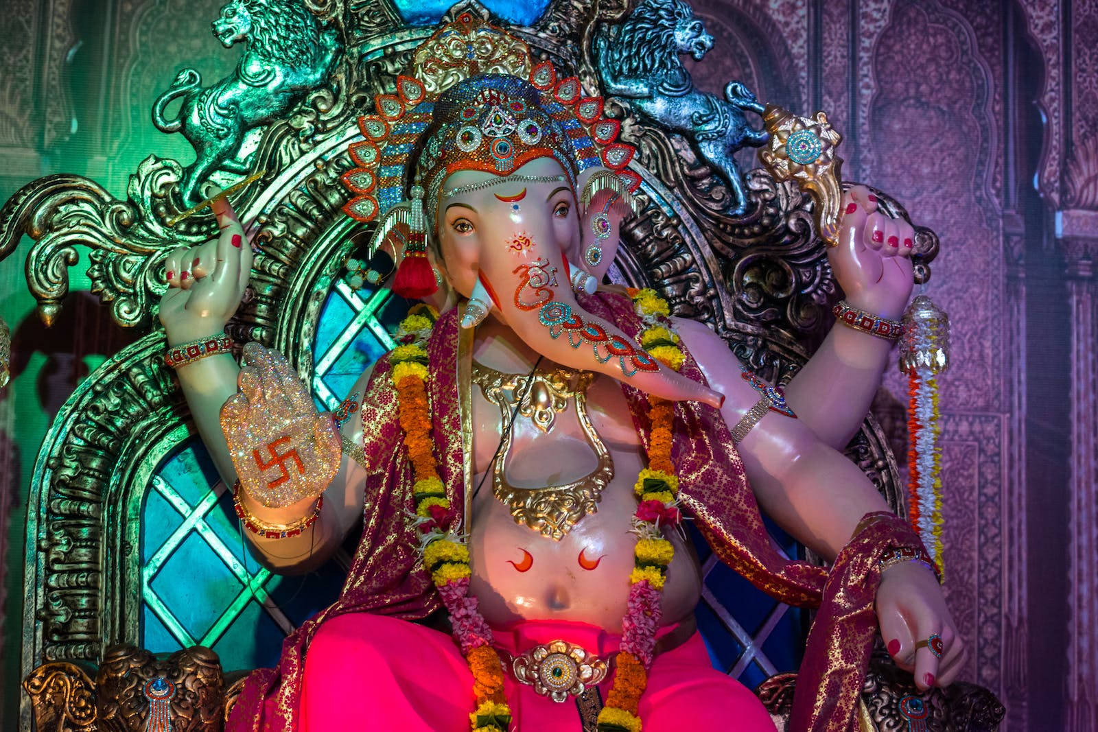 Vinayaka 桌布 壁紙 — 一尊光芒四射的象神像，身著鮮豔的粉紅色和金色服飾，戴著花環，端坐在華麗的寶座上，沐浴在多彩的燈光下。