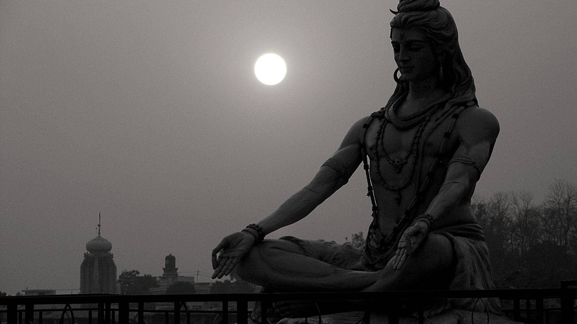 shiva dark 桌布 壁紙 — 一尊冥想神祇的黑白雕像安詳地坐在欄杆上，明亮的太陽在朦朧的城市景觀後方發光。