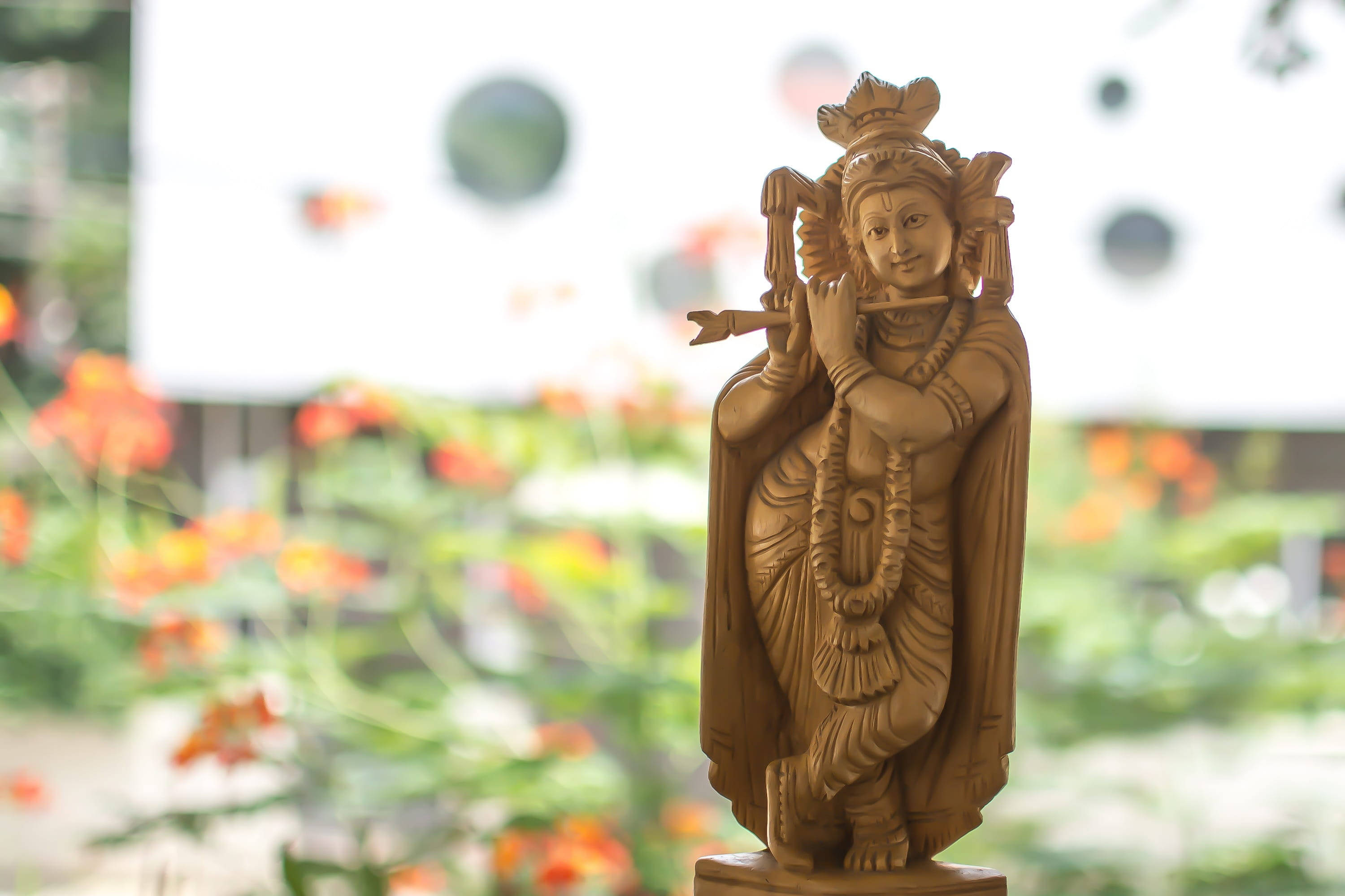 Shri Krishna 桌布 壁紙 — 一尊吹奏長笛的克里希納木雕，優雅地矗立在模糊的橙色花朵和綠葉之中。