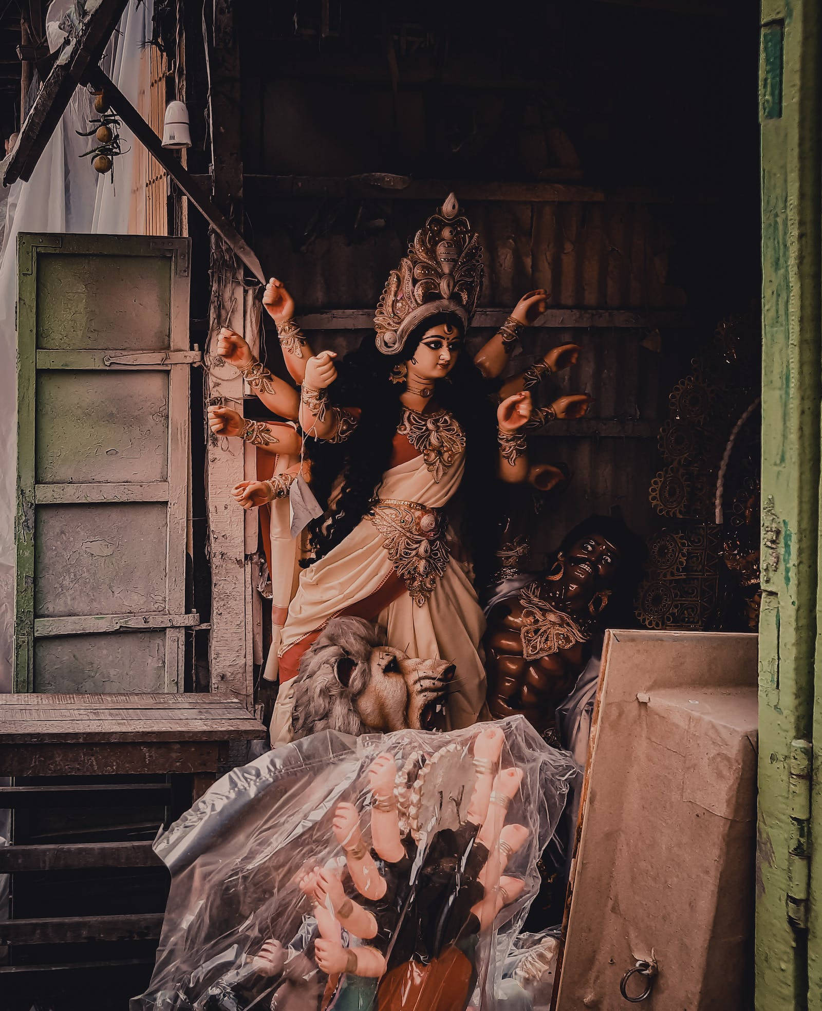maa durga 桌布 壁紙 — 一尊多臂的杜爾迦雕像莊嚴地矗立在一個雜亂的工作坊中，周圍環繞著其他神像和破舊的木結構。