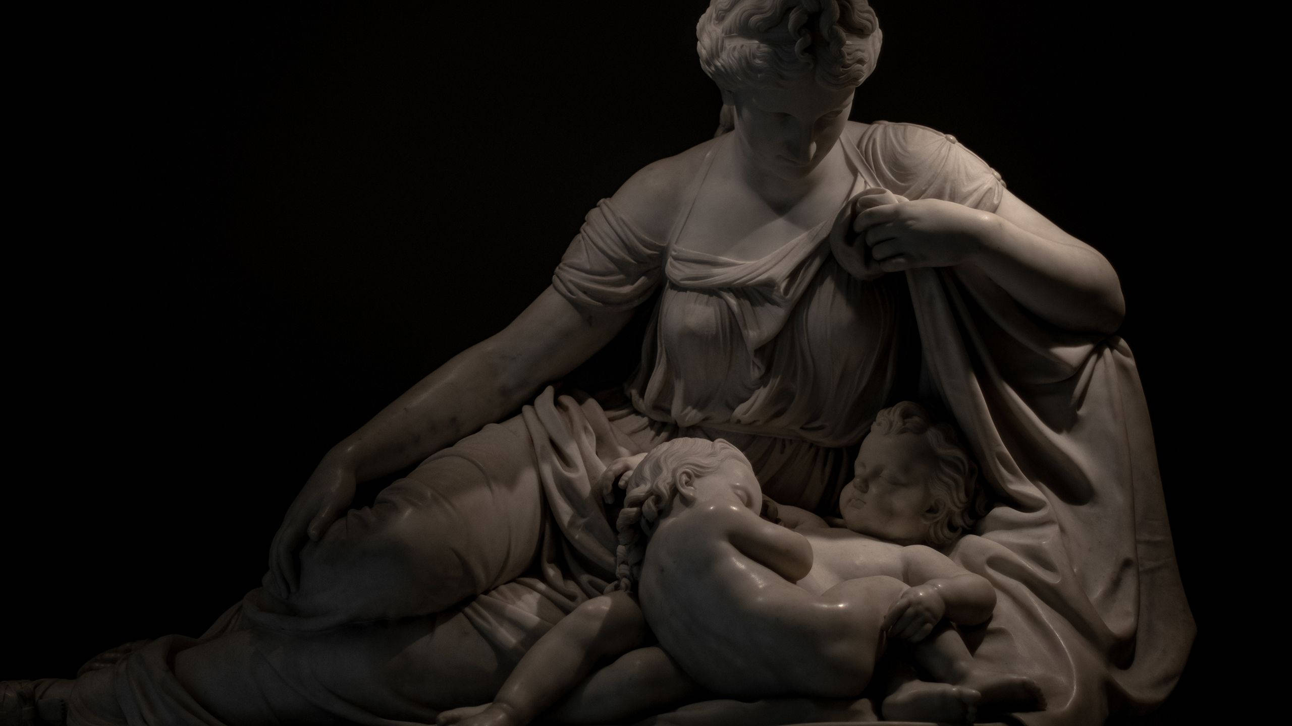 Statue Aesthetic 桌布 壁紙 — 一尊大理石雕塑，描繪一位披著布料的女性，在純黑背景下溫柔地懷抱兩個熟睡的孩子。