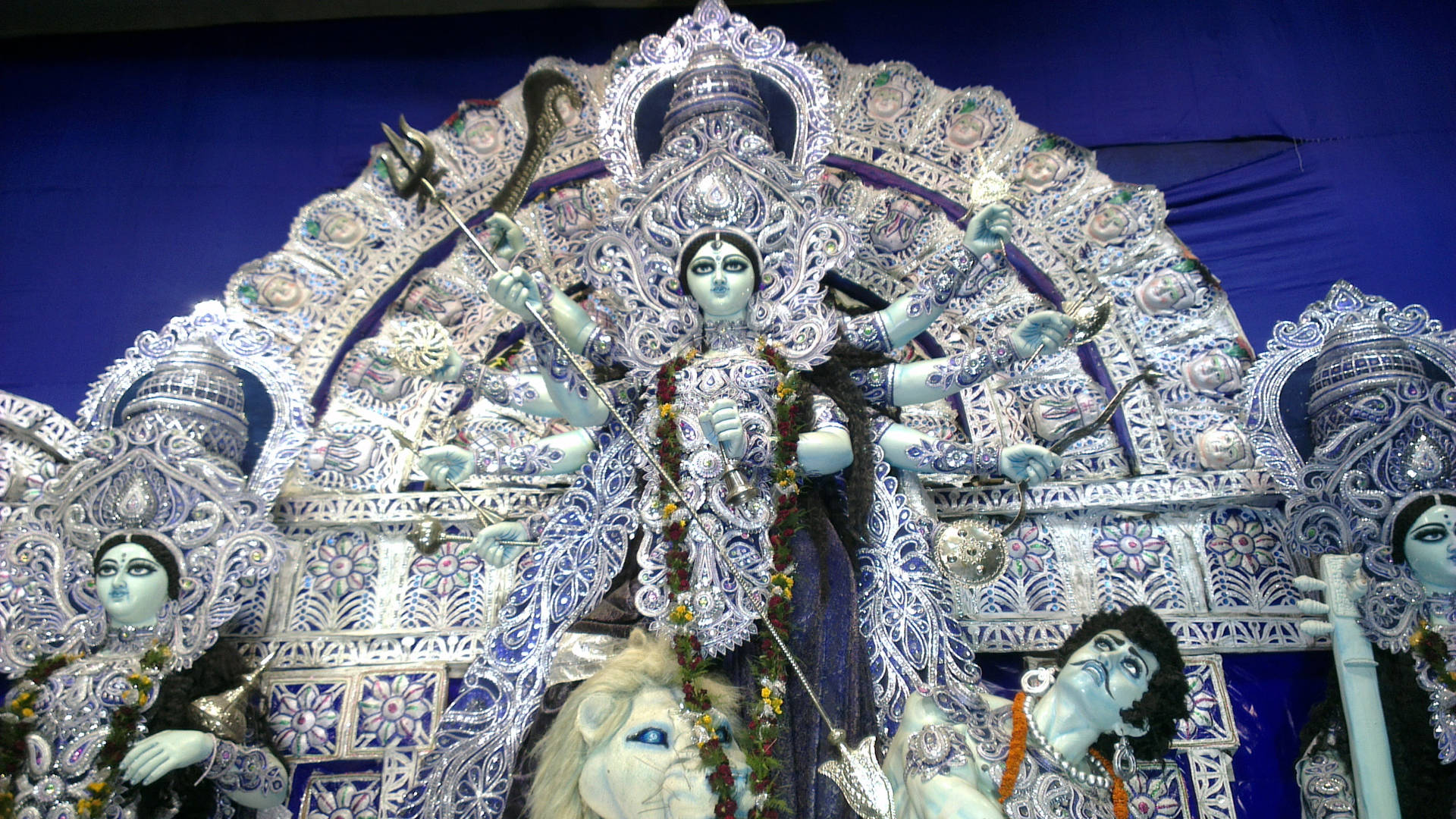 Hindu Temple 桌布 壁紙 — 一尊宏偉華麗的杜爾迦雕像，有多隻手臂和武器，立於裝飾華麗的寶座上，兩側有較小的人物。