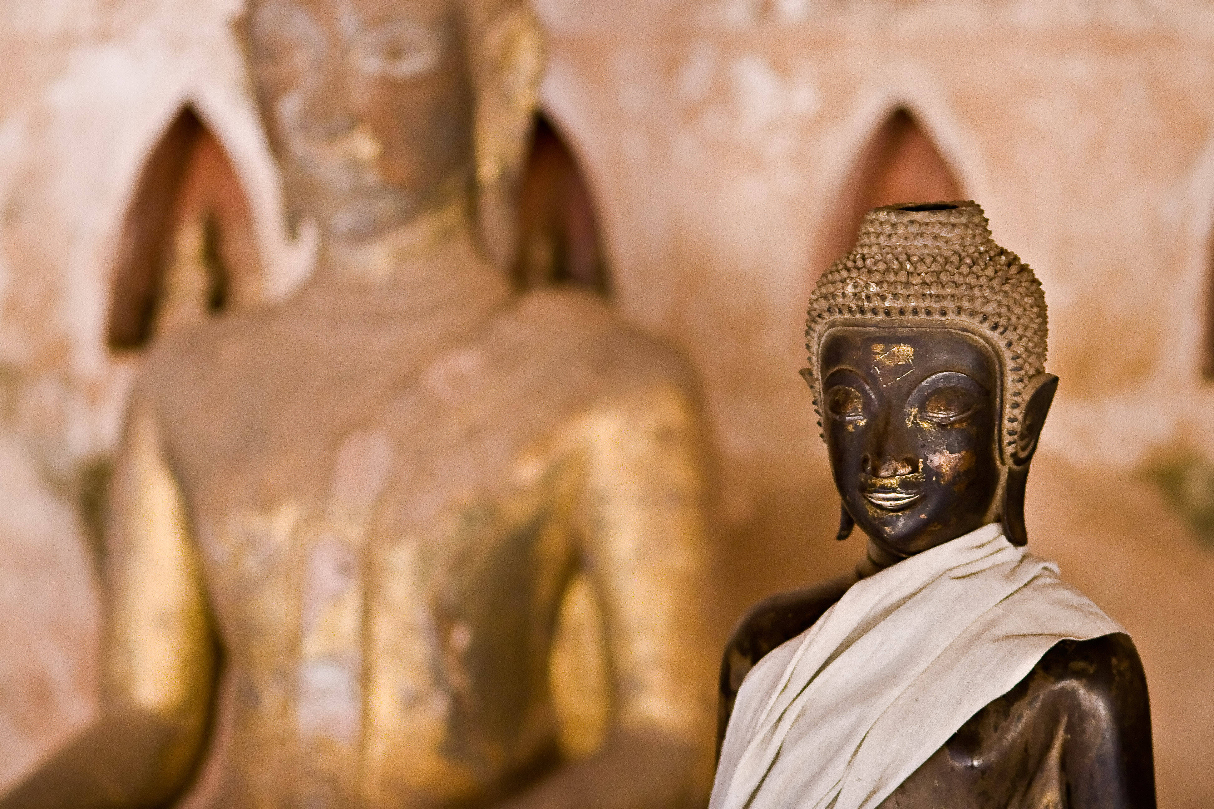 Laos 桌布 壁紙 — 一尊寧靜的深色青銅佛像披著白布立於前景，背後有模糊的金色佛像，處於溫暖的寺廟環境中。