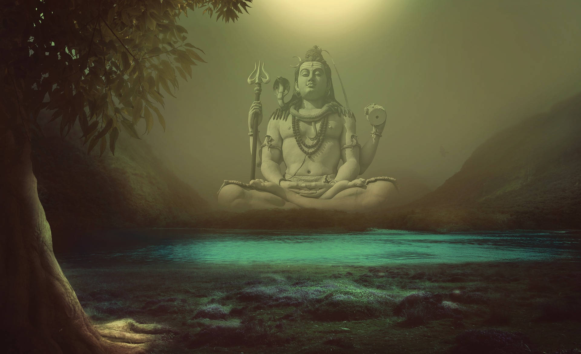 Indian 桌布 壁紙 — 一尊寧靜的濕婆石像在發光的碧綠河流上方冥想，周圍是霧氣繚繞的山脈和古樹。