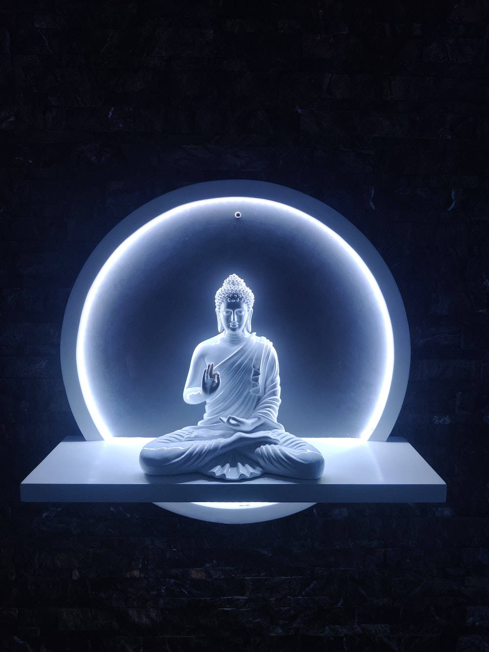 god laptop 桌布 壁紙 — 一尊寧靜的白色佛像盤腿坐在漂浮平台上，被發光的圓形光環照亮，背景是深色紋理牆壁。