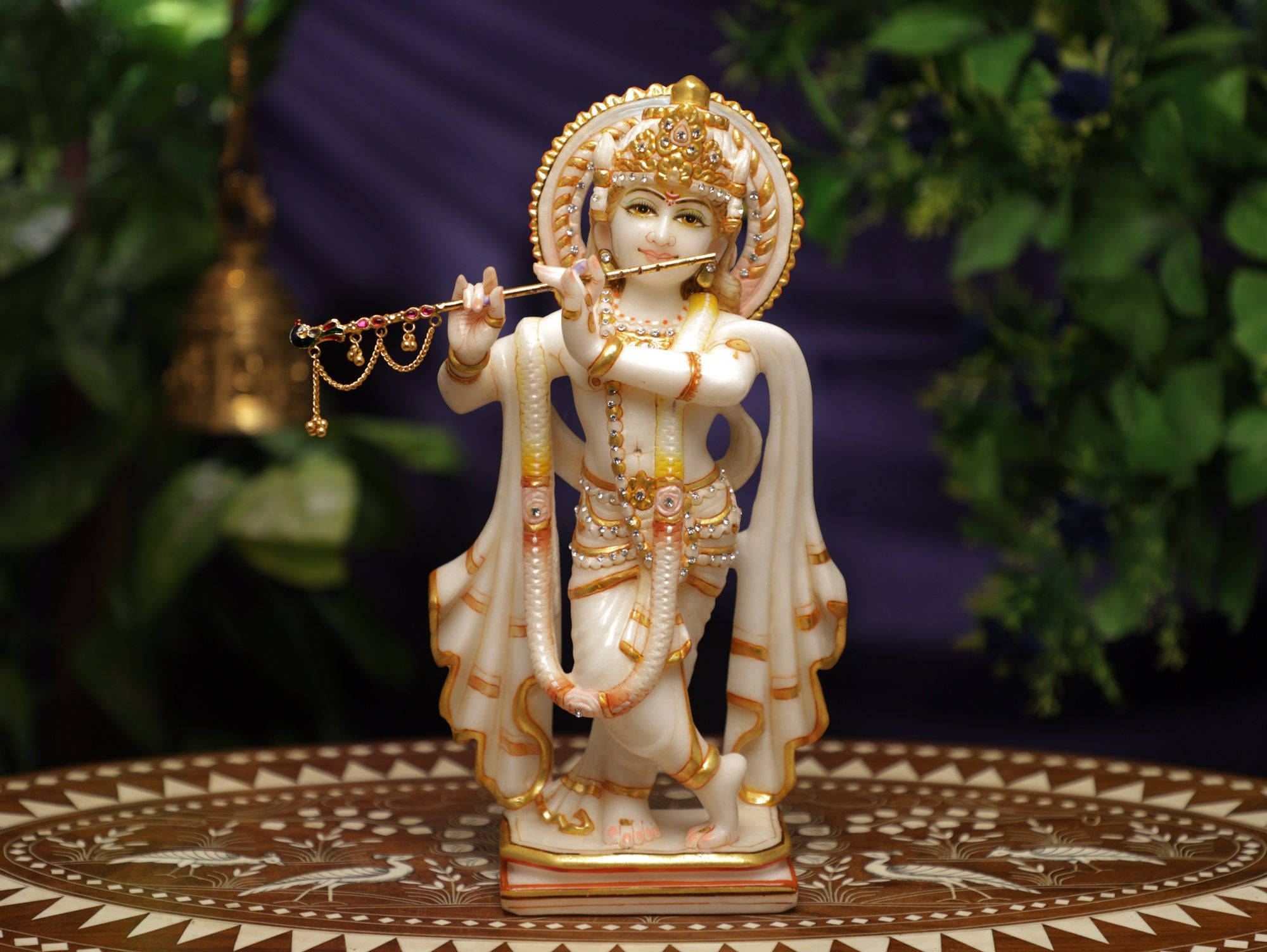 Krishna Desktop 桌布 壁紙 — 一尊寧靜的白色和金色克里希納神像正在吹奏長笛，立於一張華麗圖案桌上，背景是茂密的綠色和紫色。