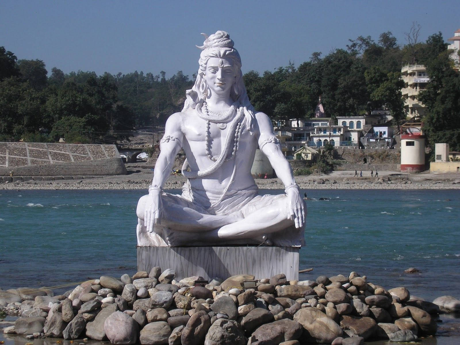 bholenath 桌布 壁紙 — 一尊寧靜的白色石雕濕婆神像在河石環繞的基座上盤腿冥想，背景有流動的河流和綠色山丘。