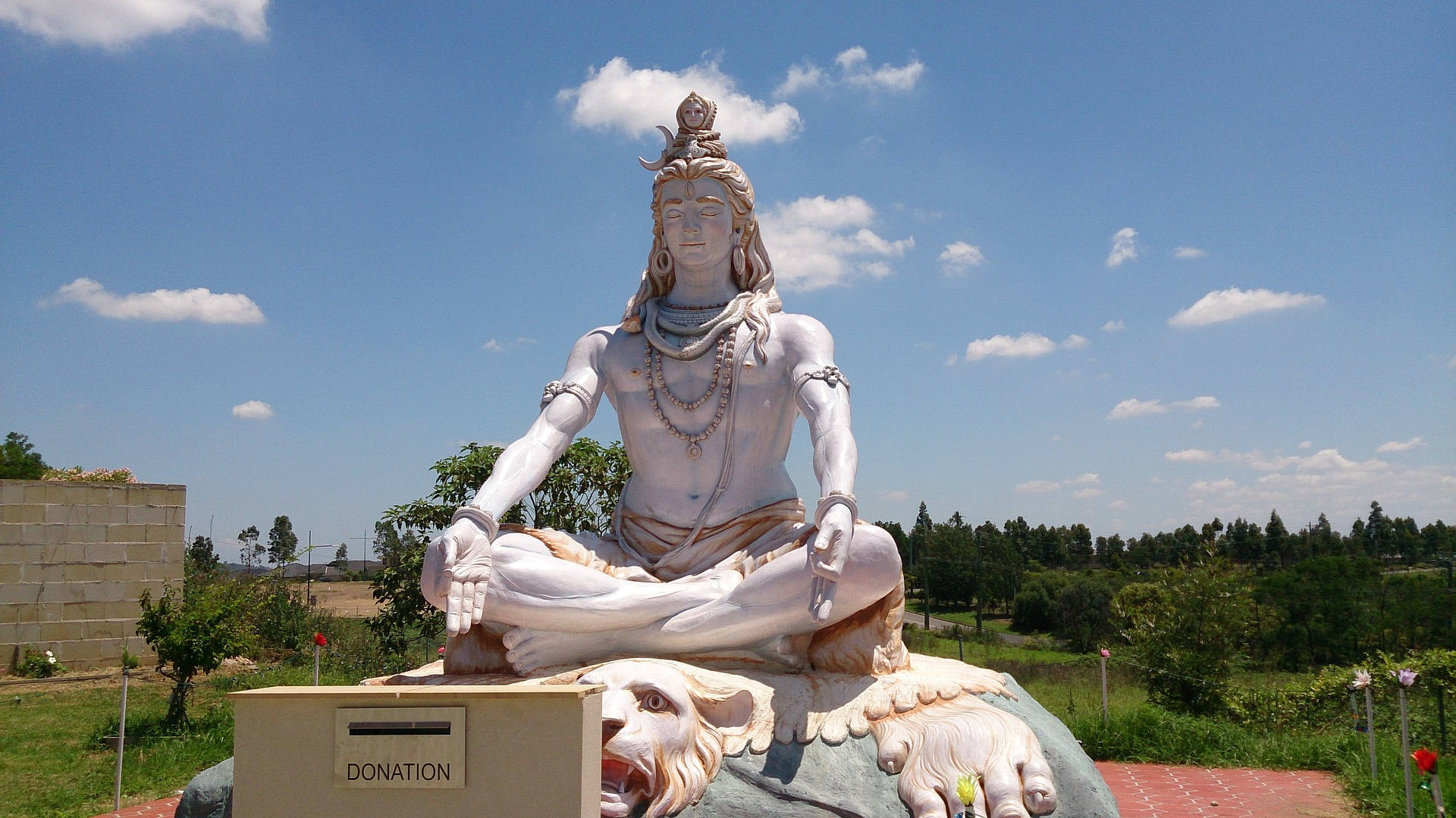 Indian 桌布 壁紙 — 一尊寧靜的白色石雕濕婆神在獅子上冥想，背景是明亮的藍天和散落的雲朵。