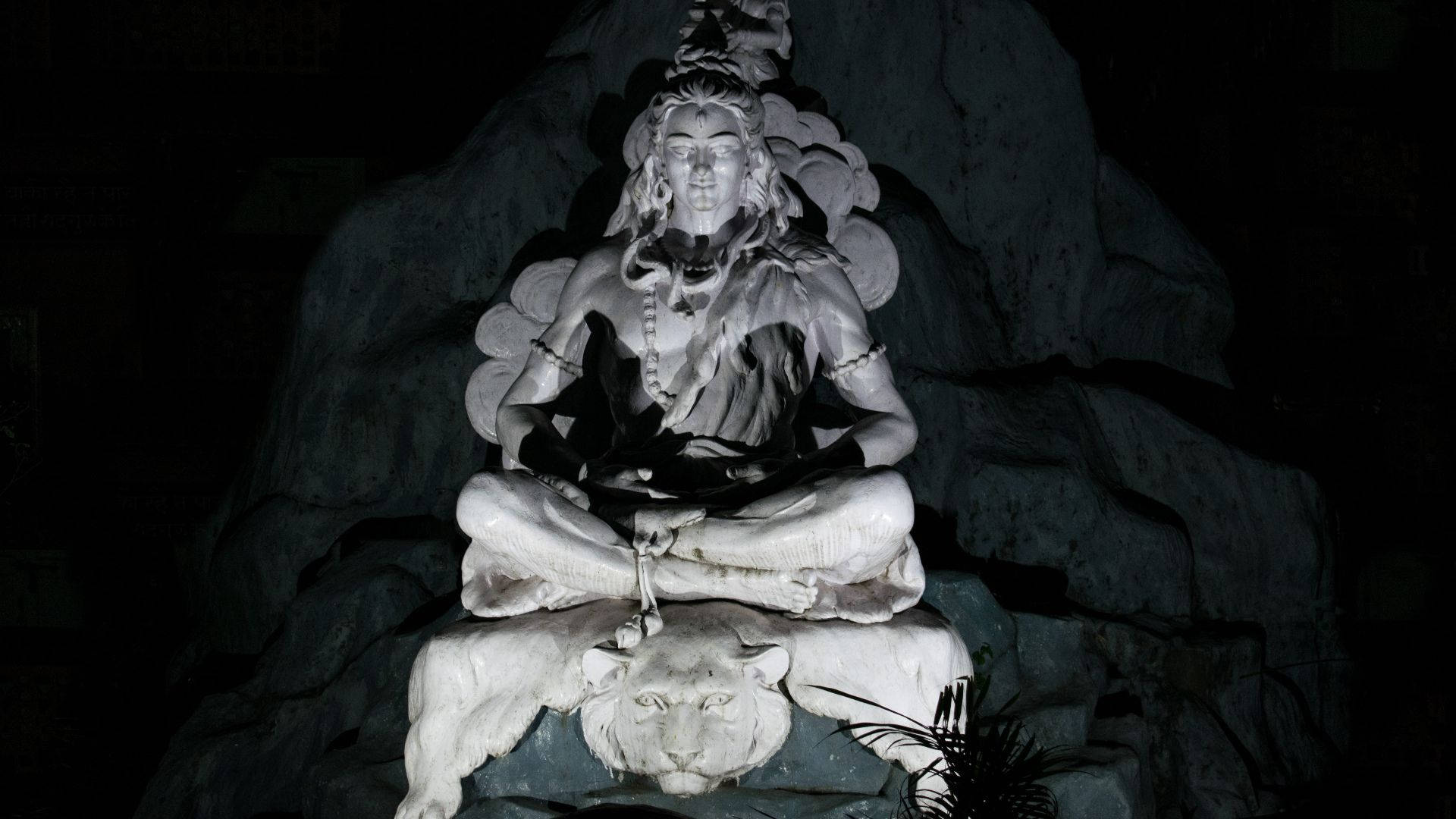 shiva dark 桌布 壁紙 — 一尊寧靜的石像，描繪一位正在冥想的菩薩，腳邊有一頭獅子，在黑暗岩石背景中以單色照明。