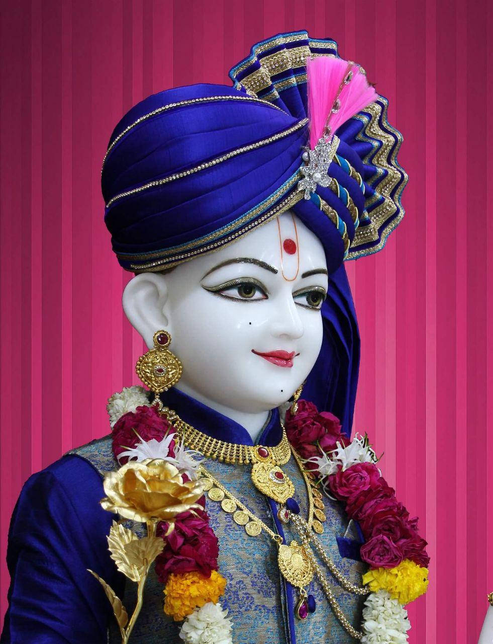 Swaminarayan 桌布 壁紙 — 一尊寧靜的神像，裝飾著皇家藍色頭巾和黃金首飾，背景是充滿活力的粉紅色條紋。