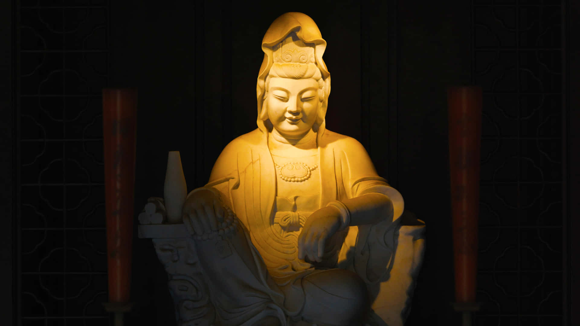 佛陀 桌布 壁紙 — 一尊寧靜的觀音石像沐浴在溫暖的金色光芒中，背景是深色圖案屏風，兩側有紅色蠟燭。