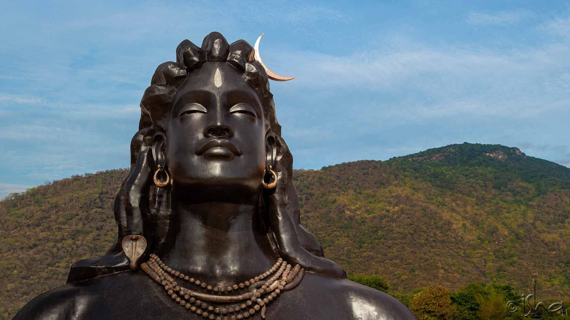 India 桌布 壁紙 — 一尊寧靜的黑色石雕濕婆神像，頭戴新月冠冕，矗立在晴朗藍天下起伏的綠色山丘背景前。