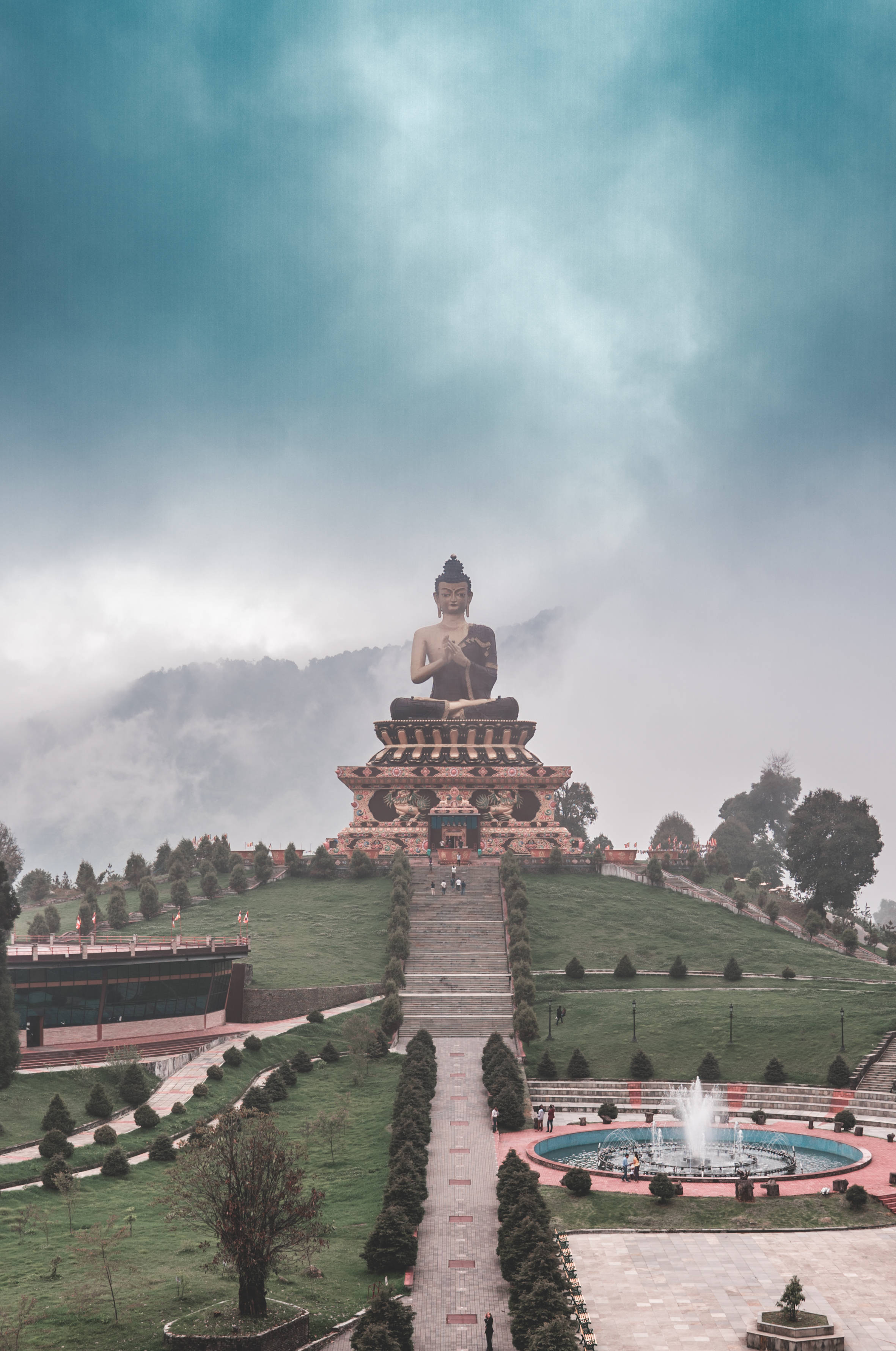 Gautam Buddha 桌布 壁紙 — 一尊巨大的佛像坐落在山頂上，周圍環繞著霧氣繚繞的山脈和修剪整齊的花園，天空多雲。