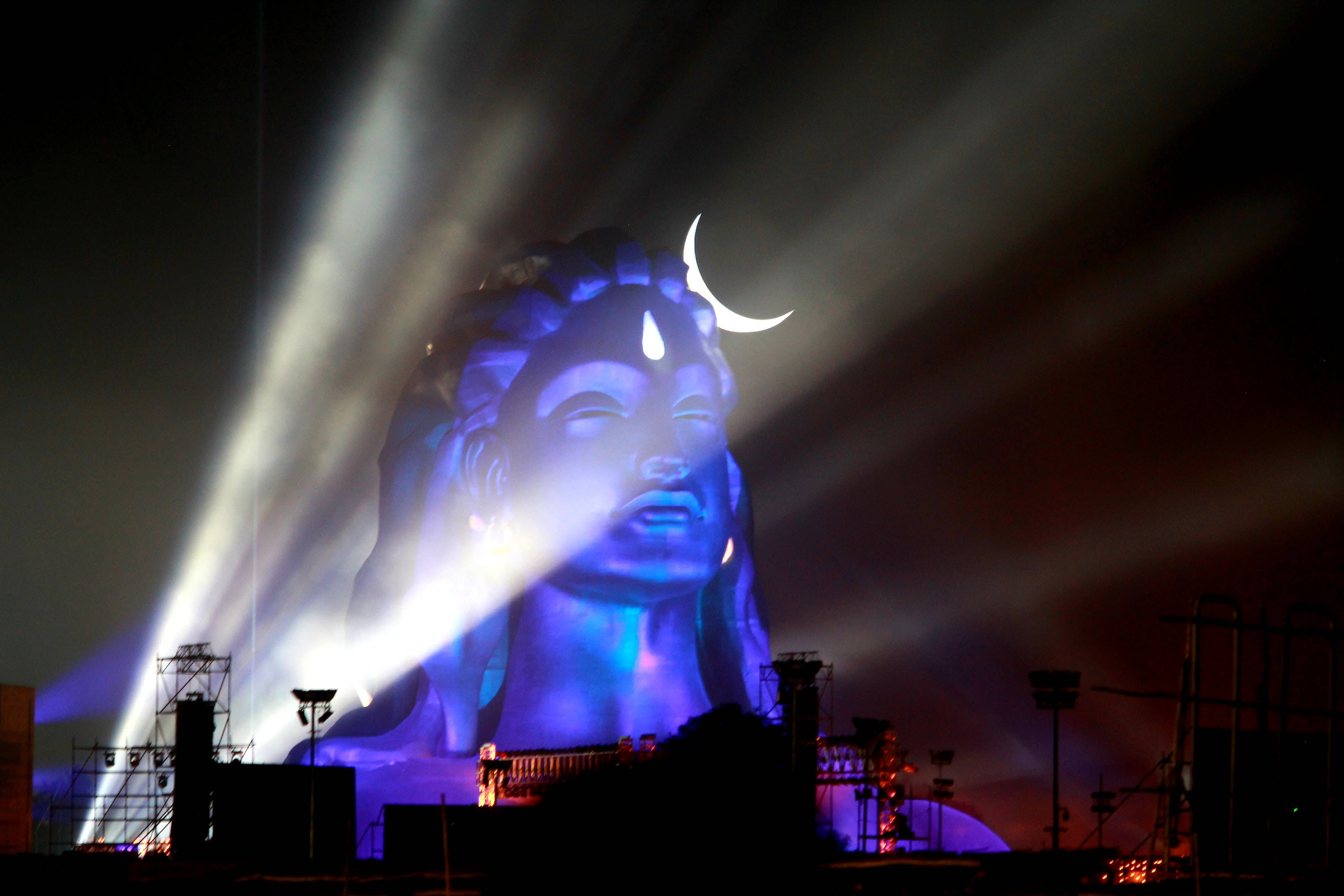 adiyogi shiva 桌布 壁紙 — 一尊巨大的佛像沐浴在藍光中，上方有一彎新月，夜間被戲劇性的聚光燈照亮。