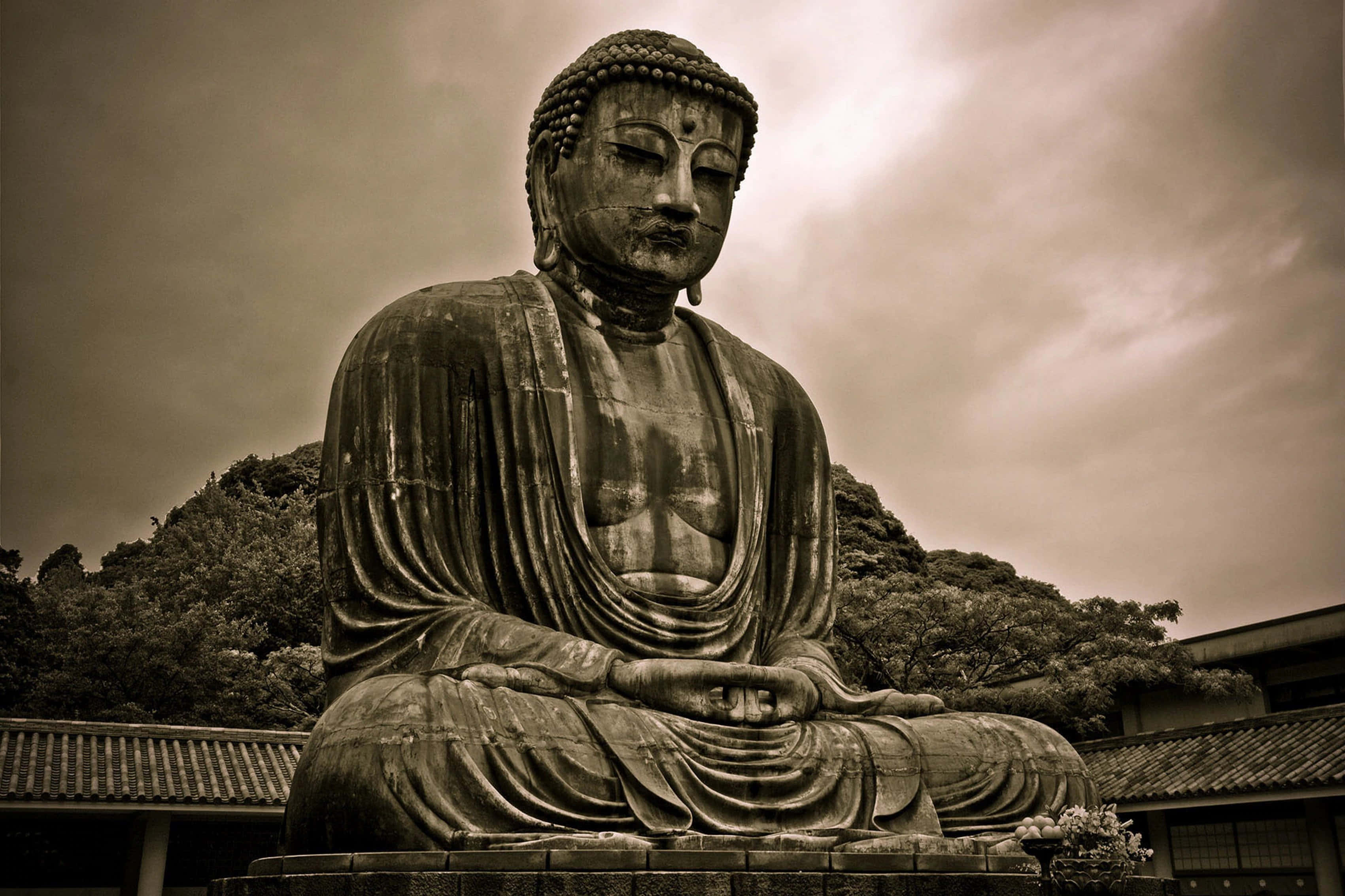Gautama Buddha 桌布 壁紙 — 一尊巨大的寧靜佛像在陰鬱的棕褐色天空下冥想，背景有樹木和傳統屋頂。