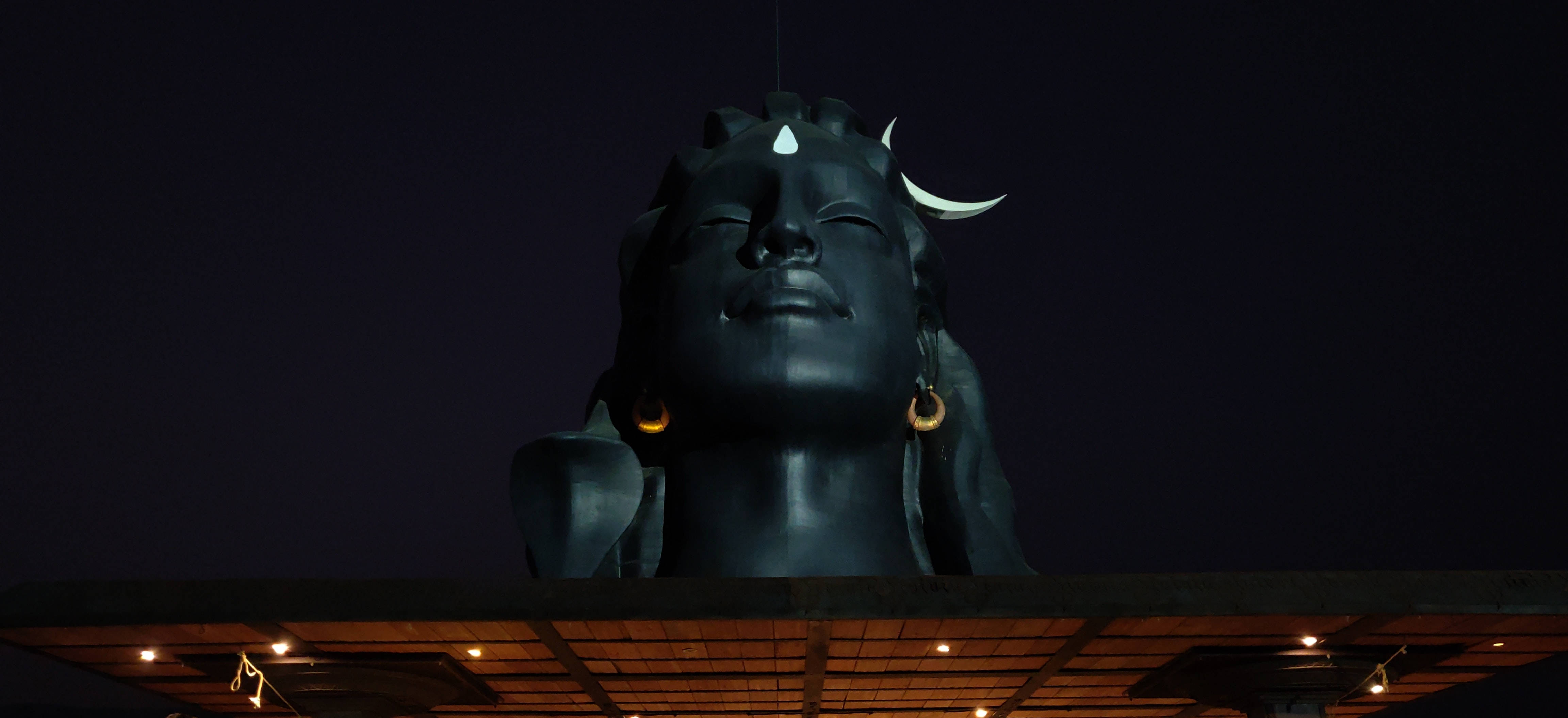 adiyogi shiva 桌布 壁紙 — 一尊巨大的深色濕婆神像，帶有新月和第三隻眼，在夜燈下對著黑色天空發光。