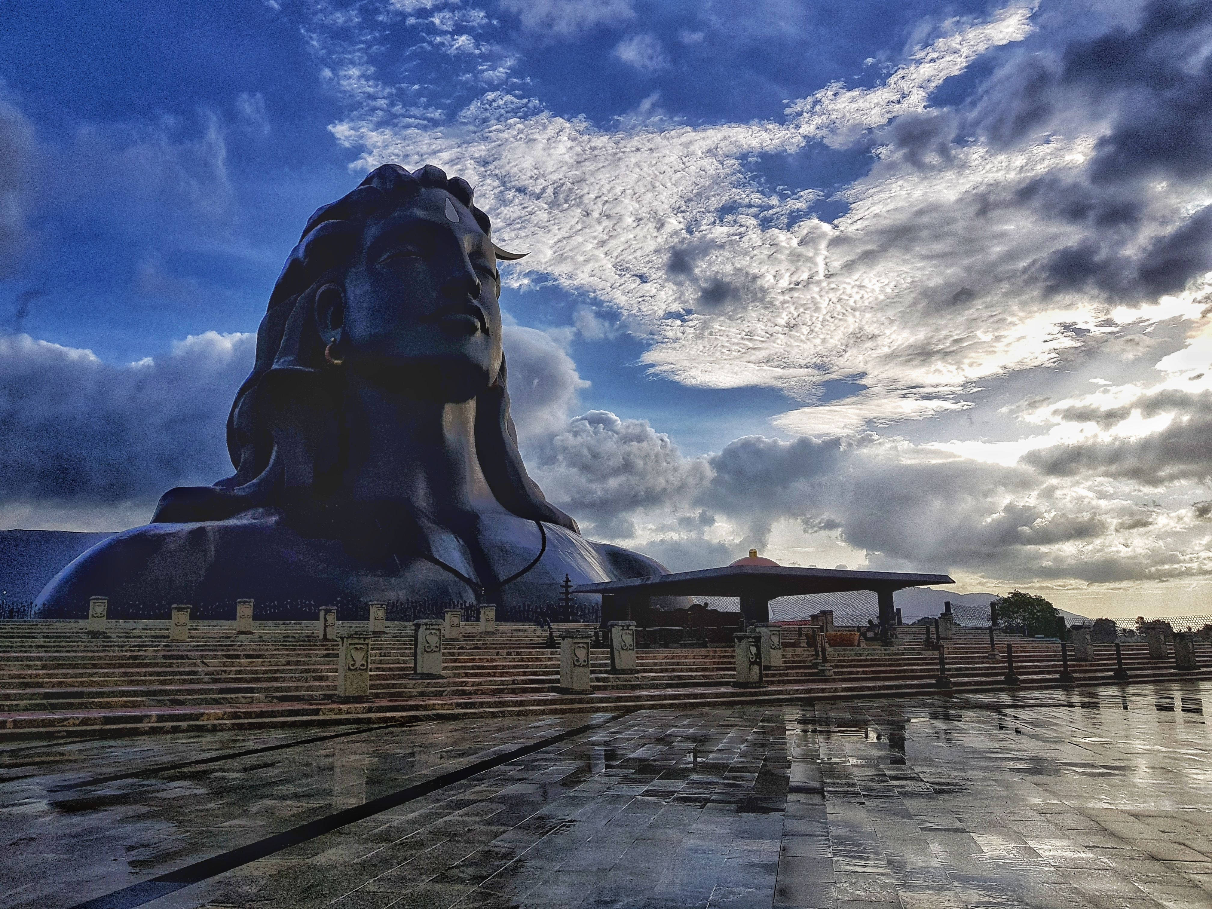 adiyogi shiva 桌布 壁紙 — 一尊巨大的深色濕婆神像，表情平靜，矗立在黃昏時分戲劇性的多雲天空下。