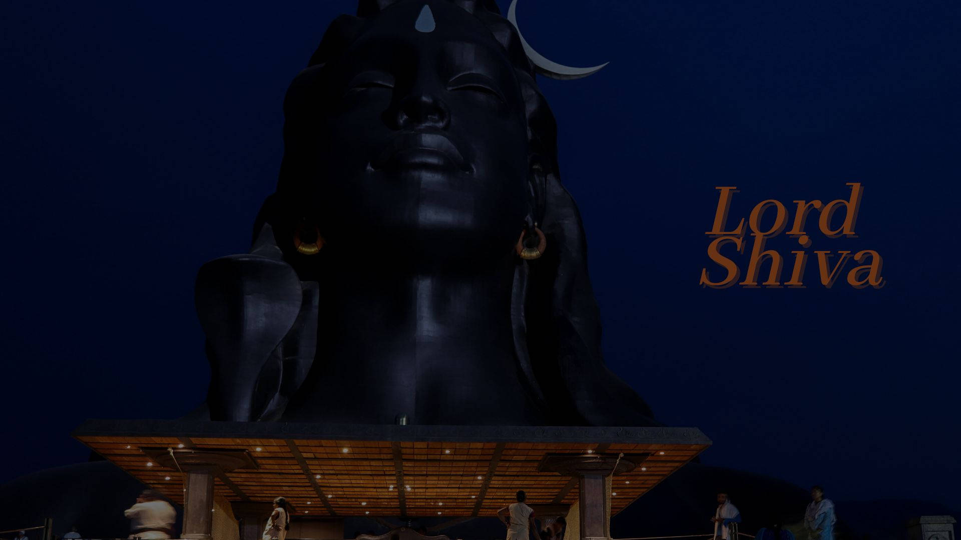 shiva dark 桌布 壁紙 — 一尊巨大的深色濕婆神像，頭戴新月冠冕，戴著金色耳環，在夜晚的深藍色天空下被照亮。