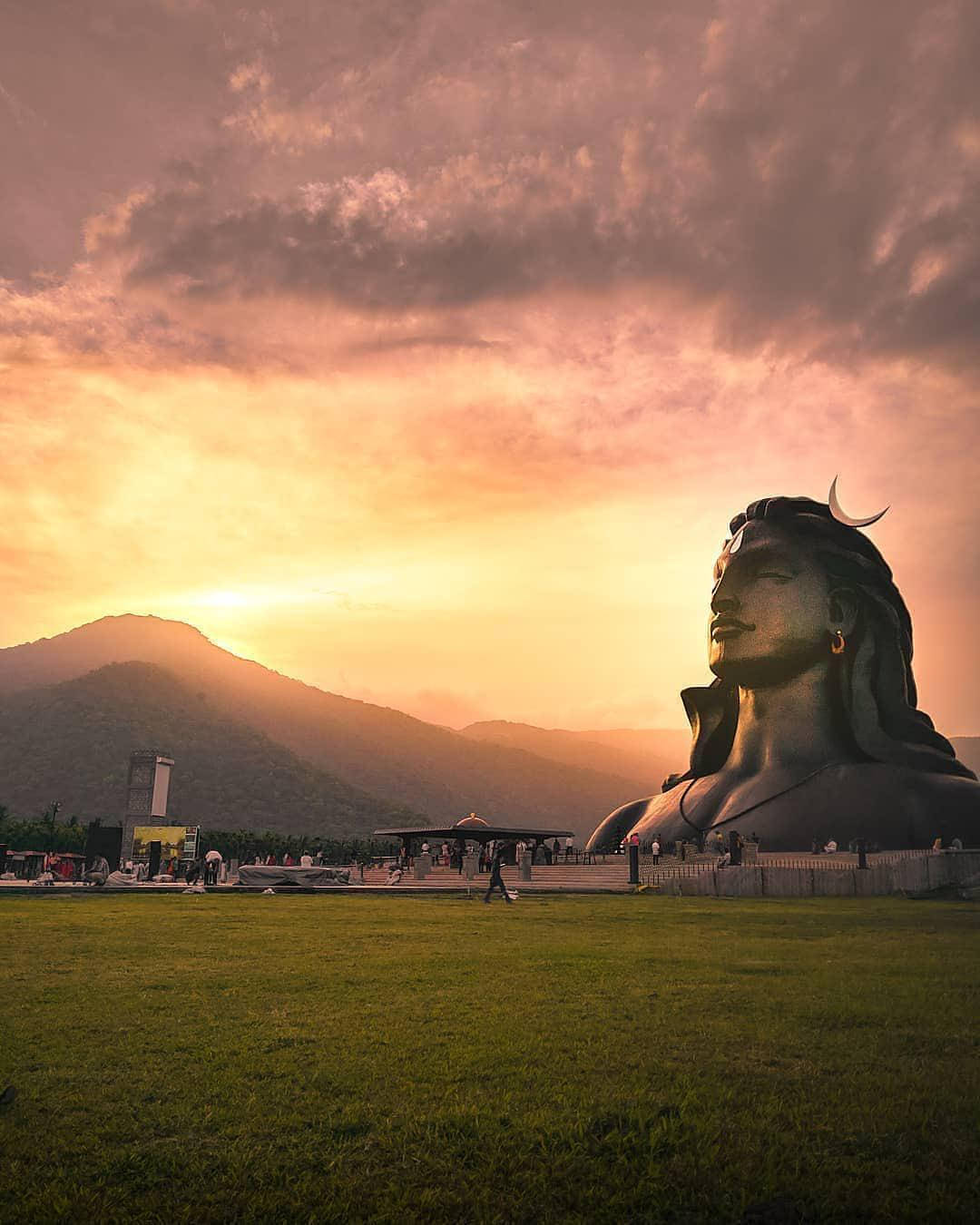 adiyogi shiva 桌布 壁紙 — 一尊巨大的濕婆神像，飾有新月，矗立在日落時分的草地上，背後山巒剪影映襯著火紅的橙色天空。