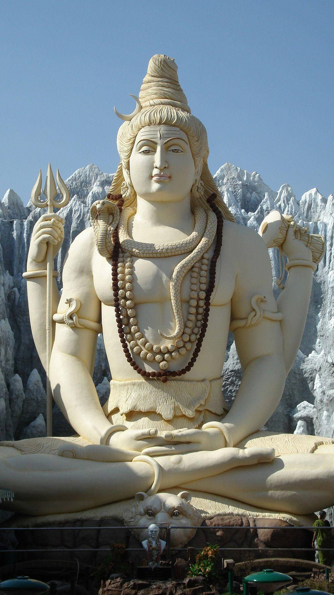 shiva iPhone 桌布 壁紙 — 一尊巨大的白色石雕濕婆神像，手持三叉戟和蛇正在冥想，背景是晴朗藍天下的人工雪山。
