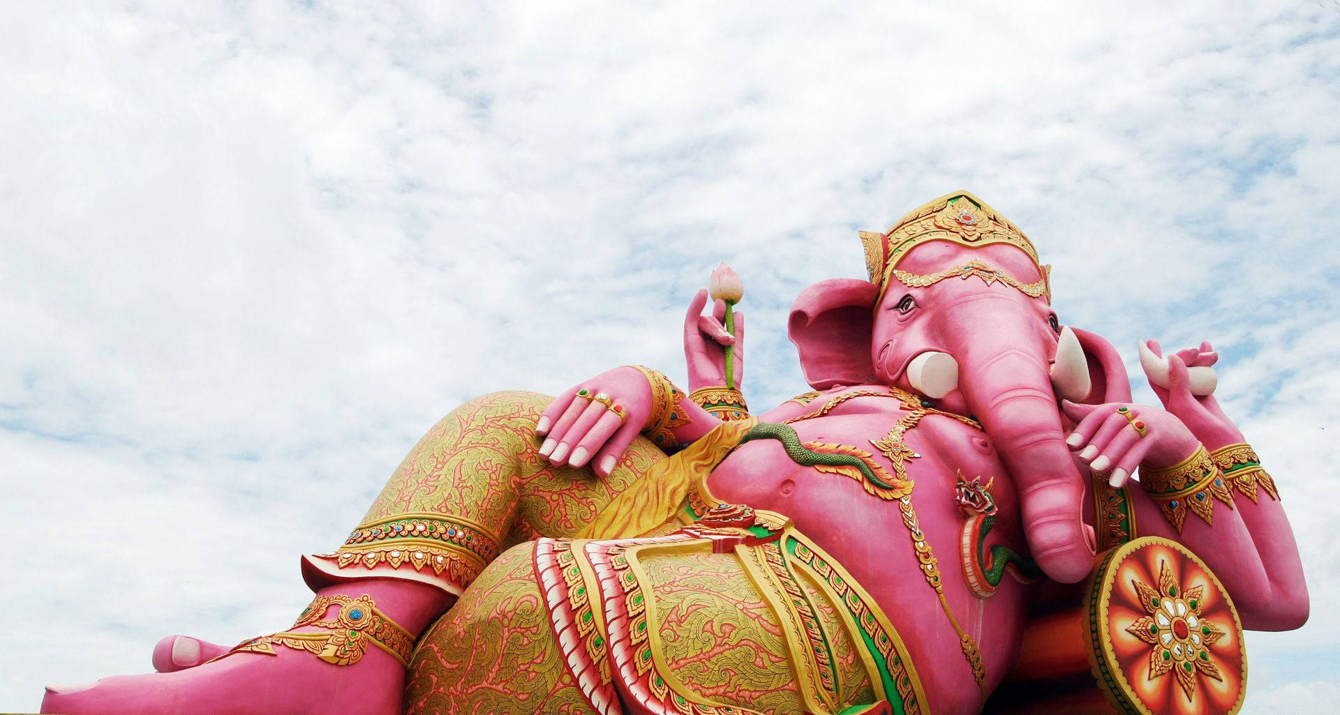 Ganesh Chaturthi 桌布 壁紙 — 一尊巨大的粉紅色象神雕像，在佈滿金色華麗細節的多雲天空下，威嚴地斜倚著。