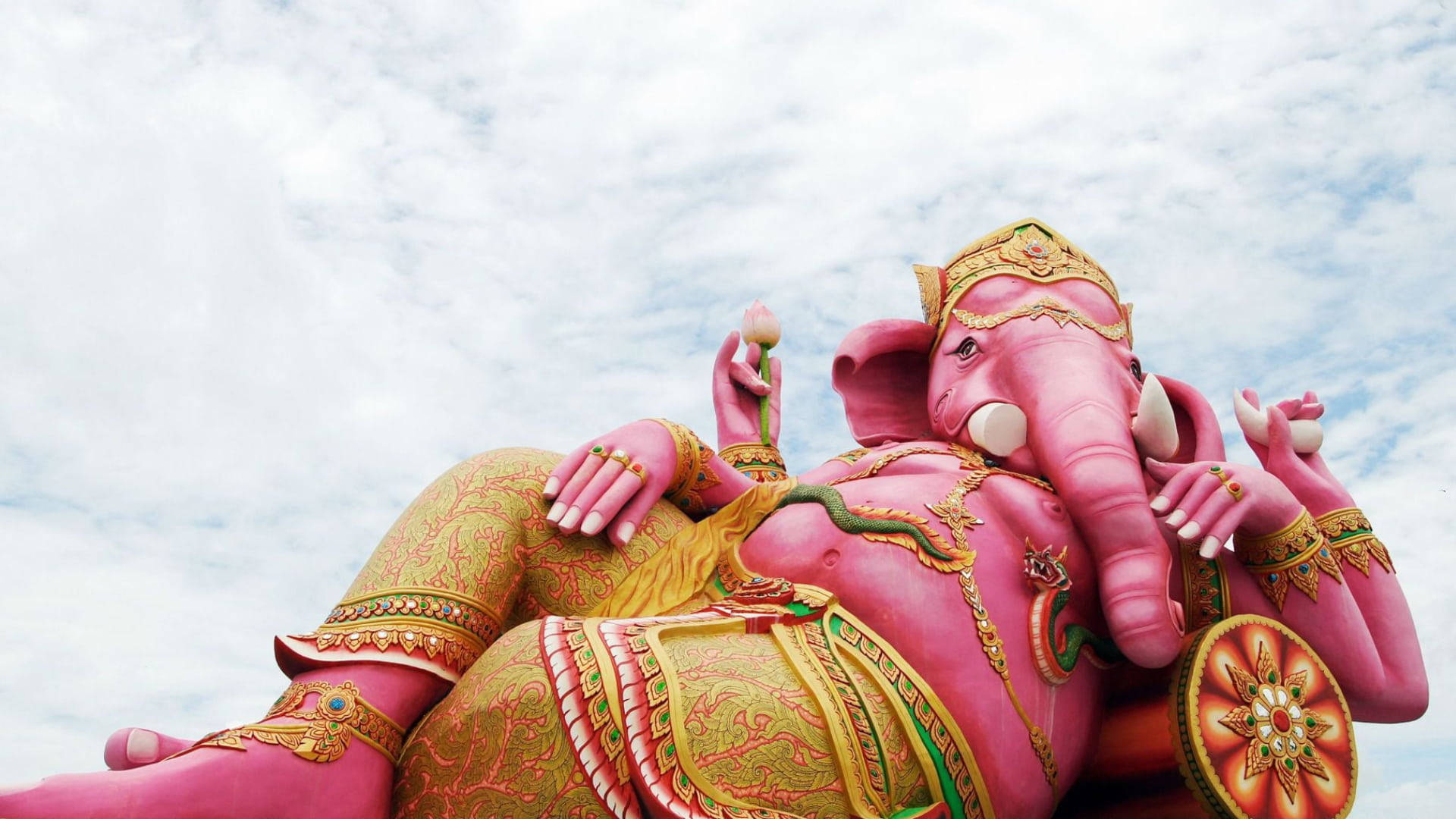 cute ganesha 桌布 壁紙 — 一尊巨大的粉紅色象頭神雕像，在點綴著金色和綠色華麗細節的多雲天空下，威嚴地斜倚著。