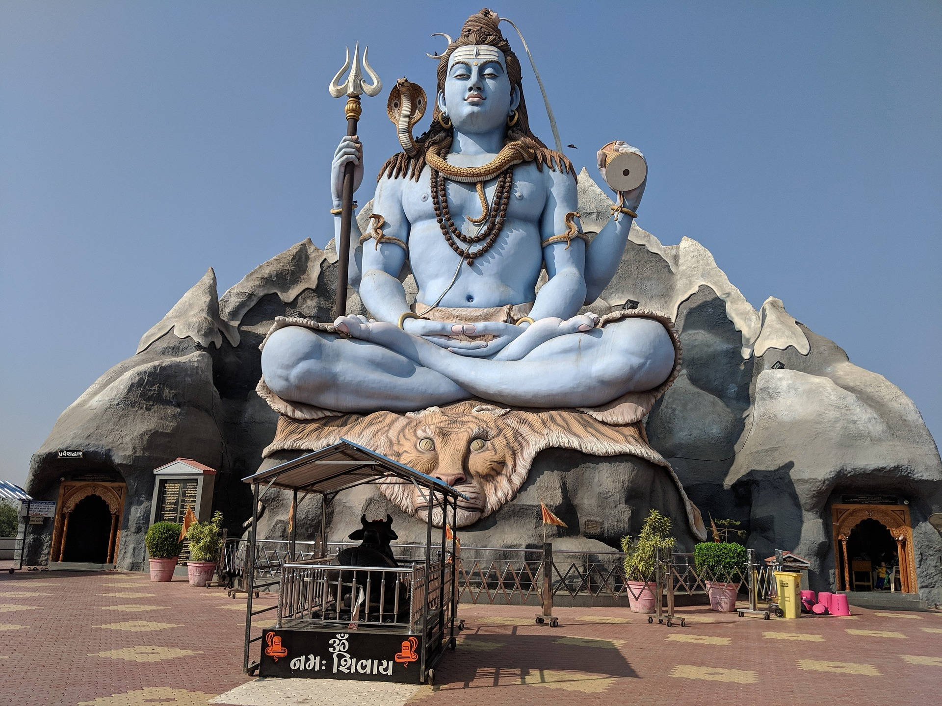 shiv shankar 桌布 壁紙 — 一尊巨大的藍色濕婆神像，在虎皮上冥想，手持三叉戟和達瑪魯，背景是晴朗的天空。