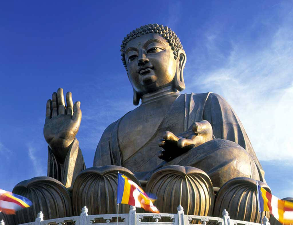 god laptop 桌布 壁紙 — 一尊巨大的金色佛像在晴朗的藍天下安詳地坐著，下方有色彩繽紛的經幡飄揚。