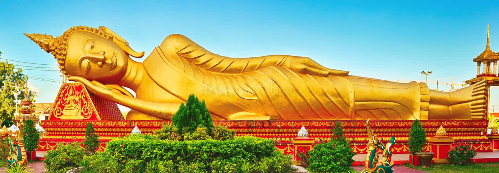 Golden 桌布 壁紙 — 一尊巨大的金色臥佛在晴朗的藍天下安詳地躺著，周圍有鮮紅色的寺廟建築和茂密的綠色植物。